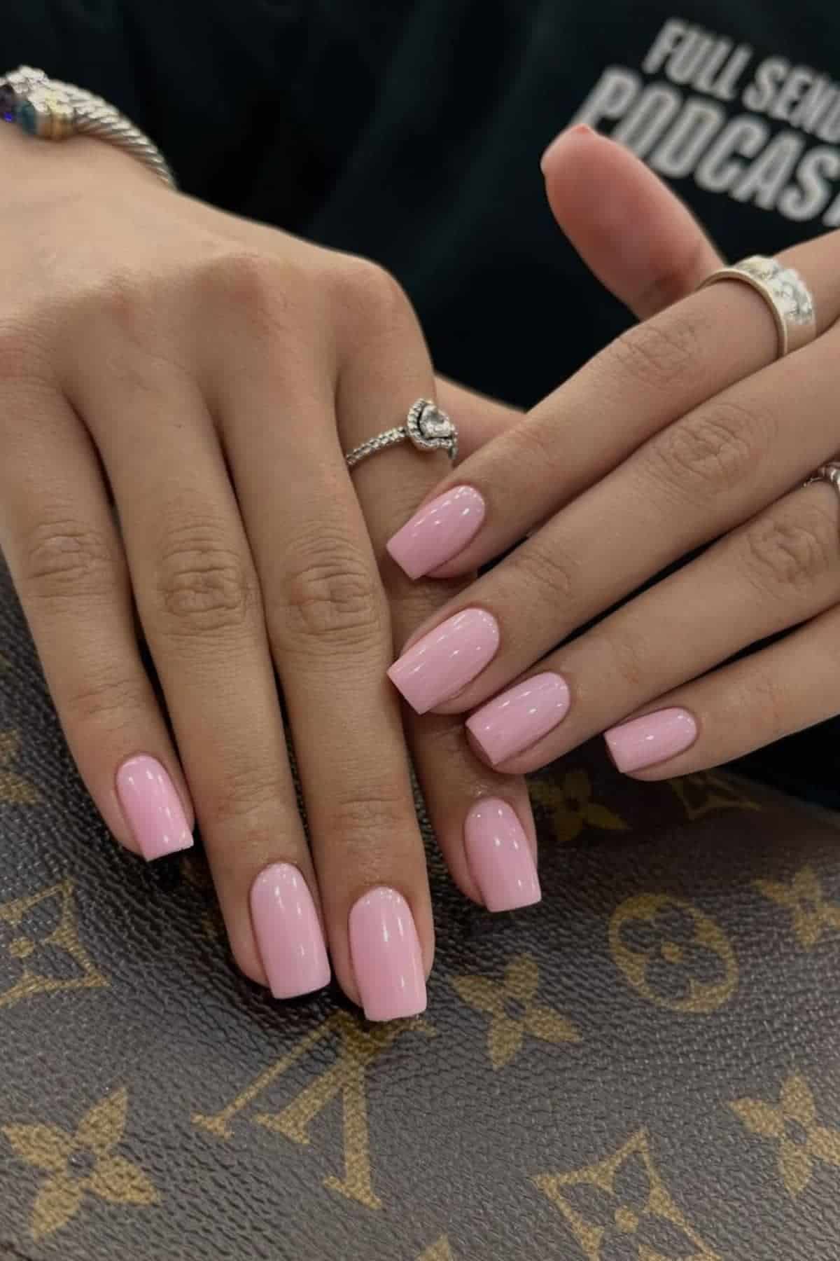 Close-up of hands with long, glossy, light pink manicured nails resting on a brown Louis Vuitton bag. The person is wearing rings and a dark top.