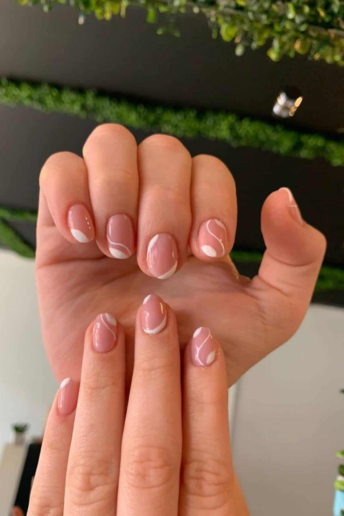 A hand with short, almond-shaped nails featuring a nude base and minimal white swirl designs, held up against a background with green foliage.