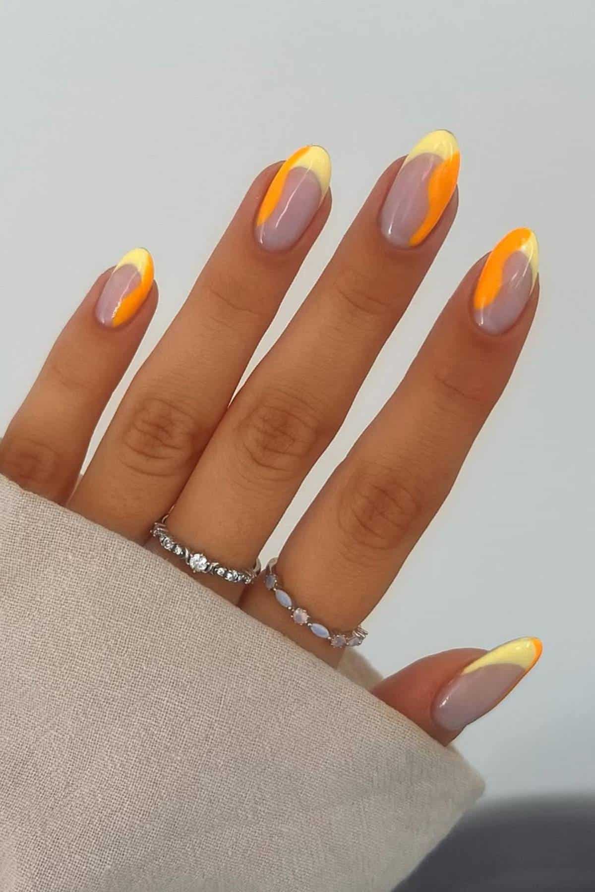 A hand with almond-shaped nails painted in a gradient design featuring yellow, orange, and mauve colors. The person is wearing silver rings and a beige long-sleeve top.