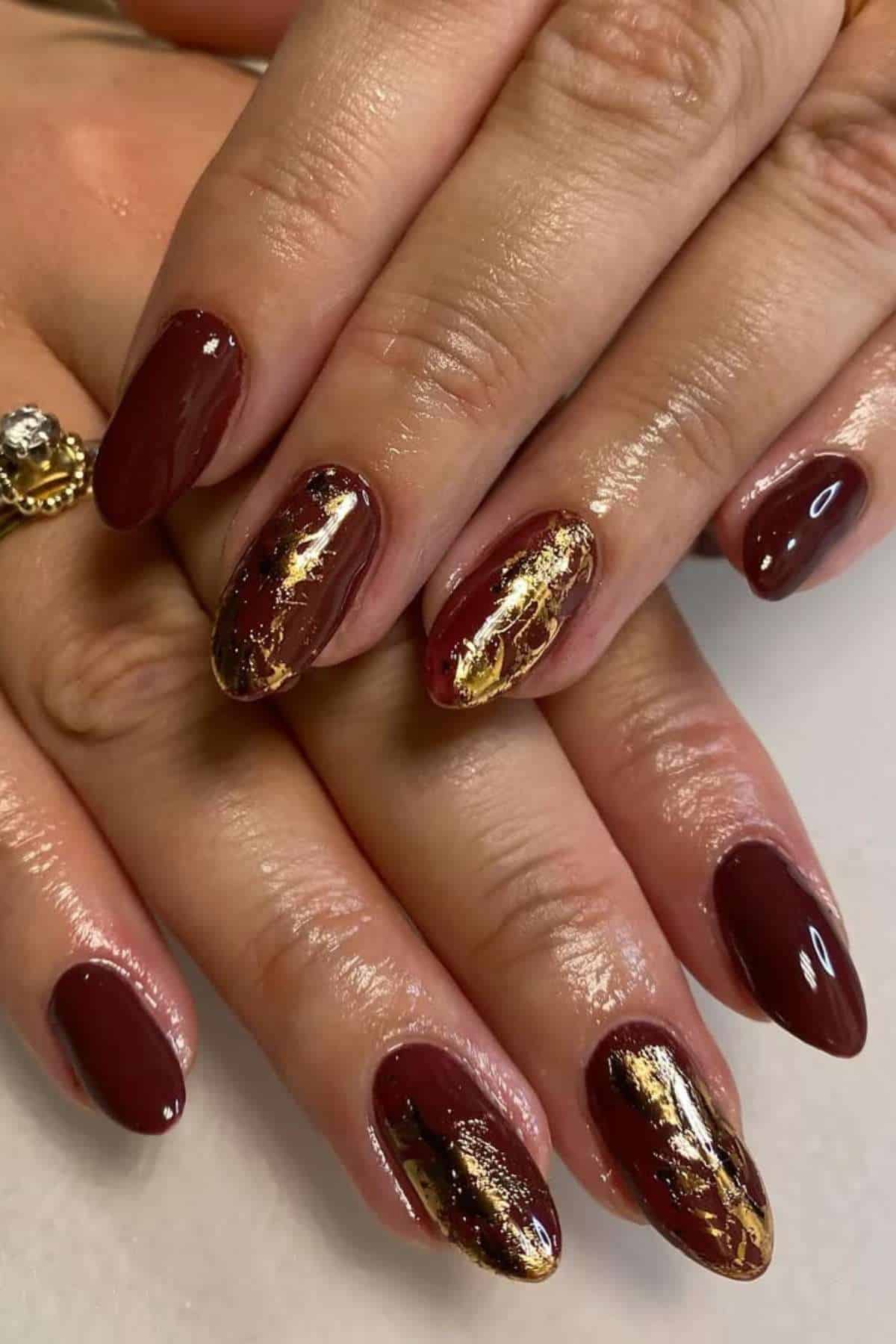 Hands with oval-shaped maroon nail polish; two accent nails on each hand feature gold foil detailing. A gold ring with a clear stone is on one finger.