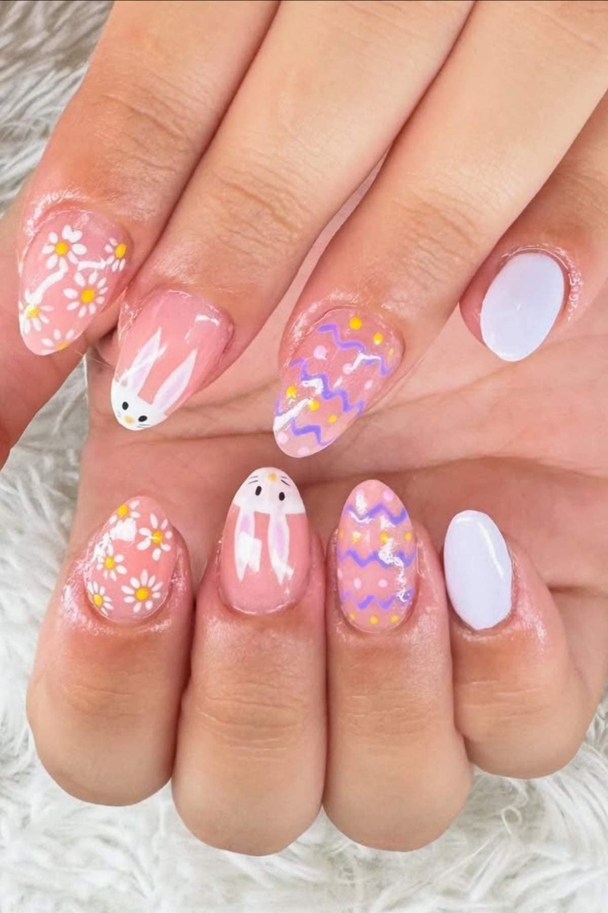 Hands with pastel-colored Easter-themed nail art, including daisies, bunny faces, zigzag patterns, and solid lavender nails.