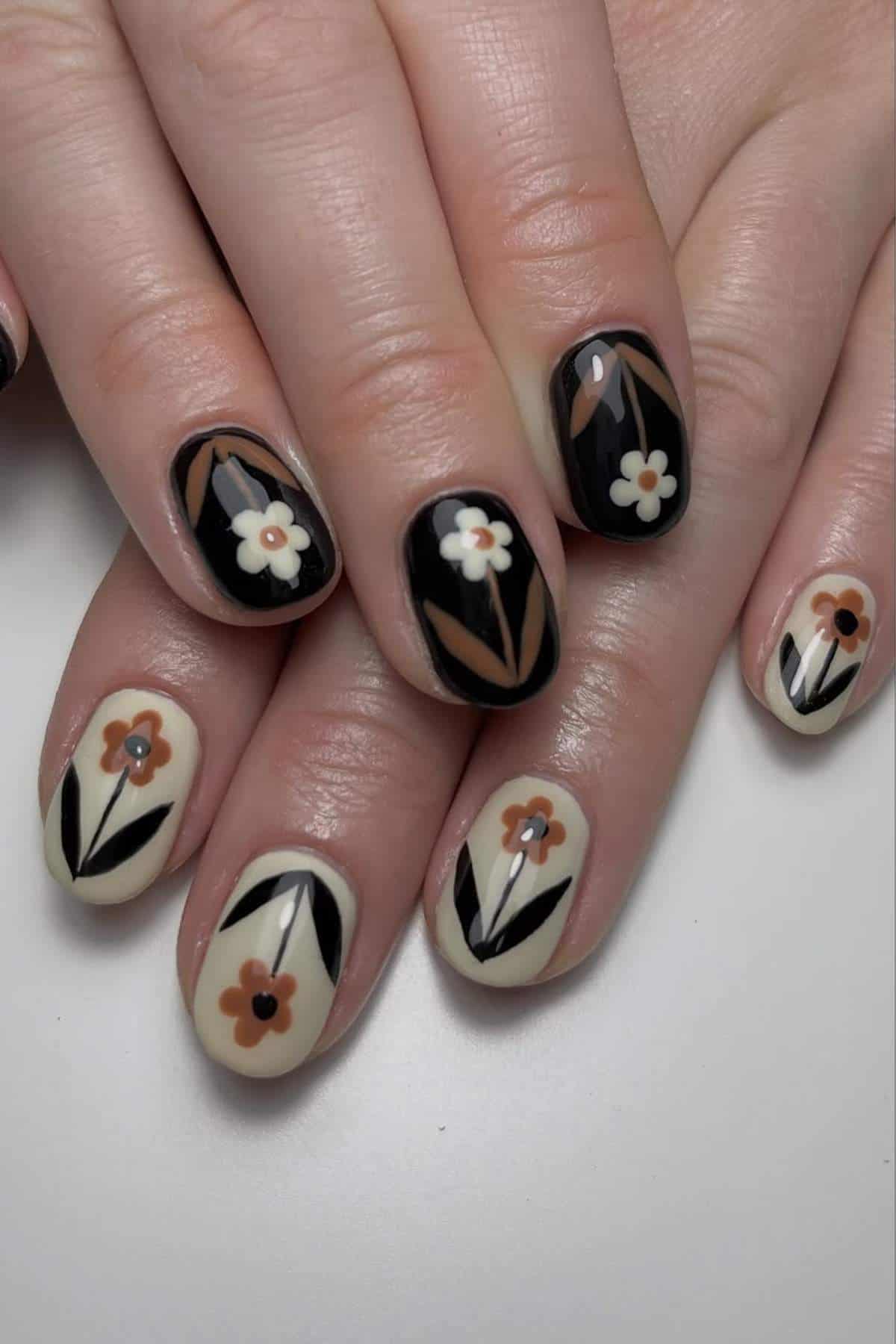 Close-up of hands with manicured nails featuring black and beige backgrounds and floral designs in white, orange, and brown.