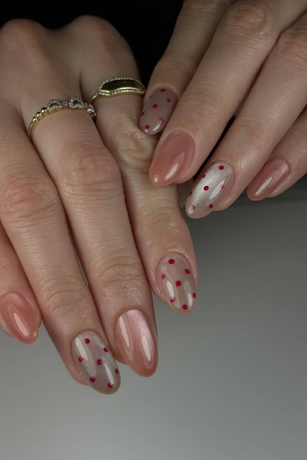 Close-up of hands with almond-shaped nails featuring a nude base color, some decorated with red polka dots. The person is wearing gold rings.