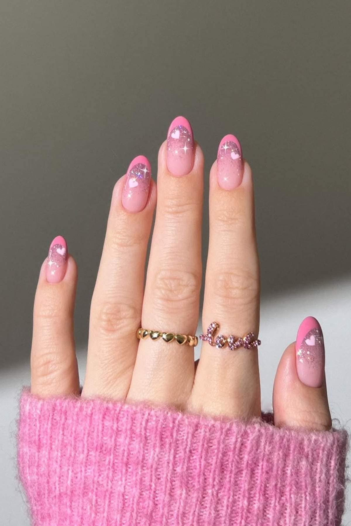 A hand with pink and white gradient manicure featuring small heart designs, wearing two gold rings, and a pink knit sleeve partially covering the wrist.