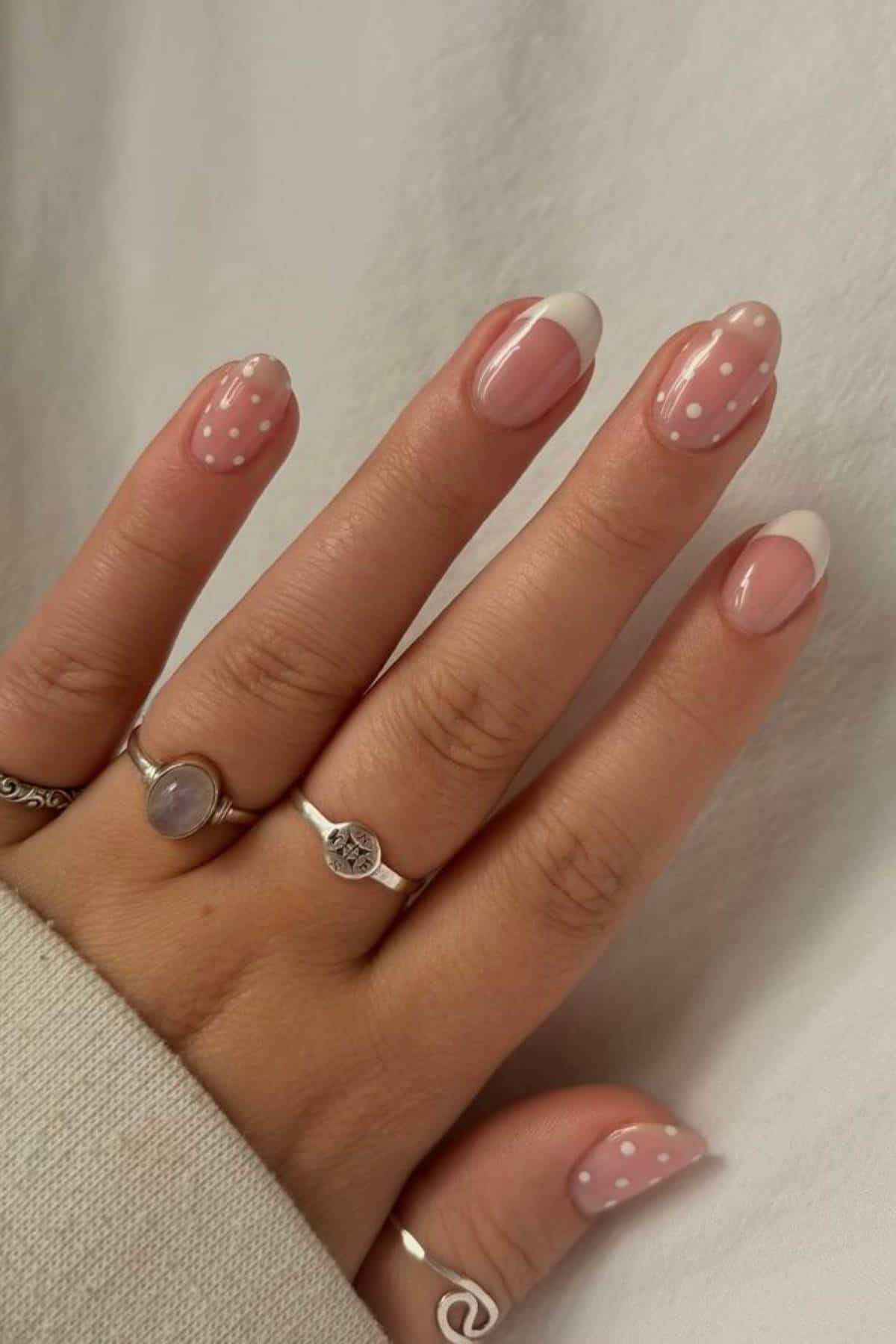 A hand with almond-shaped nails featuring a pink base, white French tips, and white polka dot designs. The person is wearing three silver rings.