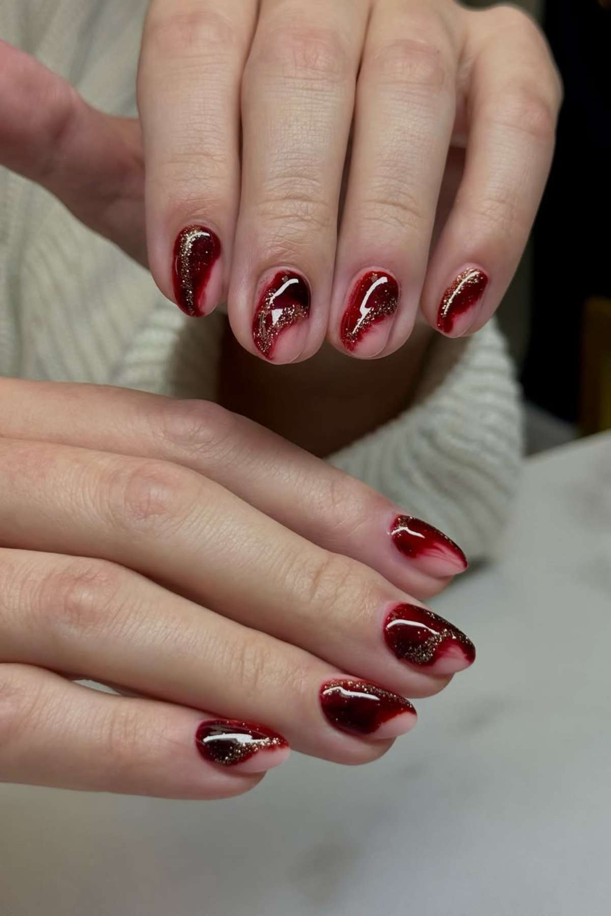 Close-up of hands with red and black ombre nails featuring gold glitter accents and abstract wavy designs.