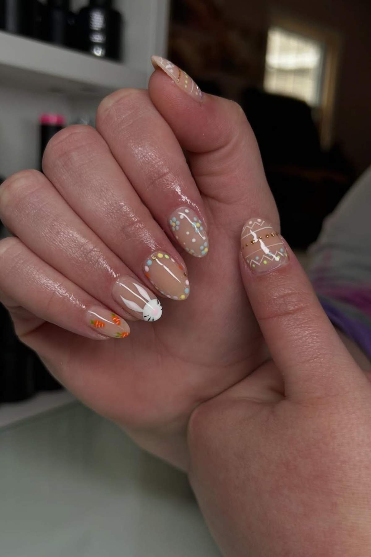 A hand with almond-shaped nails featuring beige polish and intricate white, orange, and green geometric and polka dot designs.