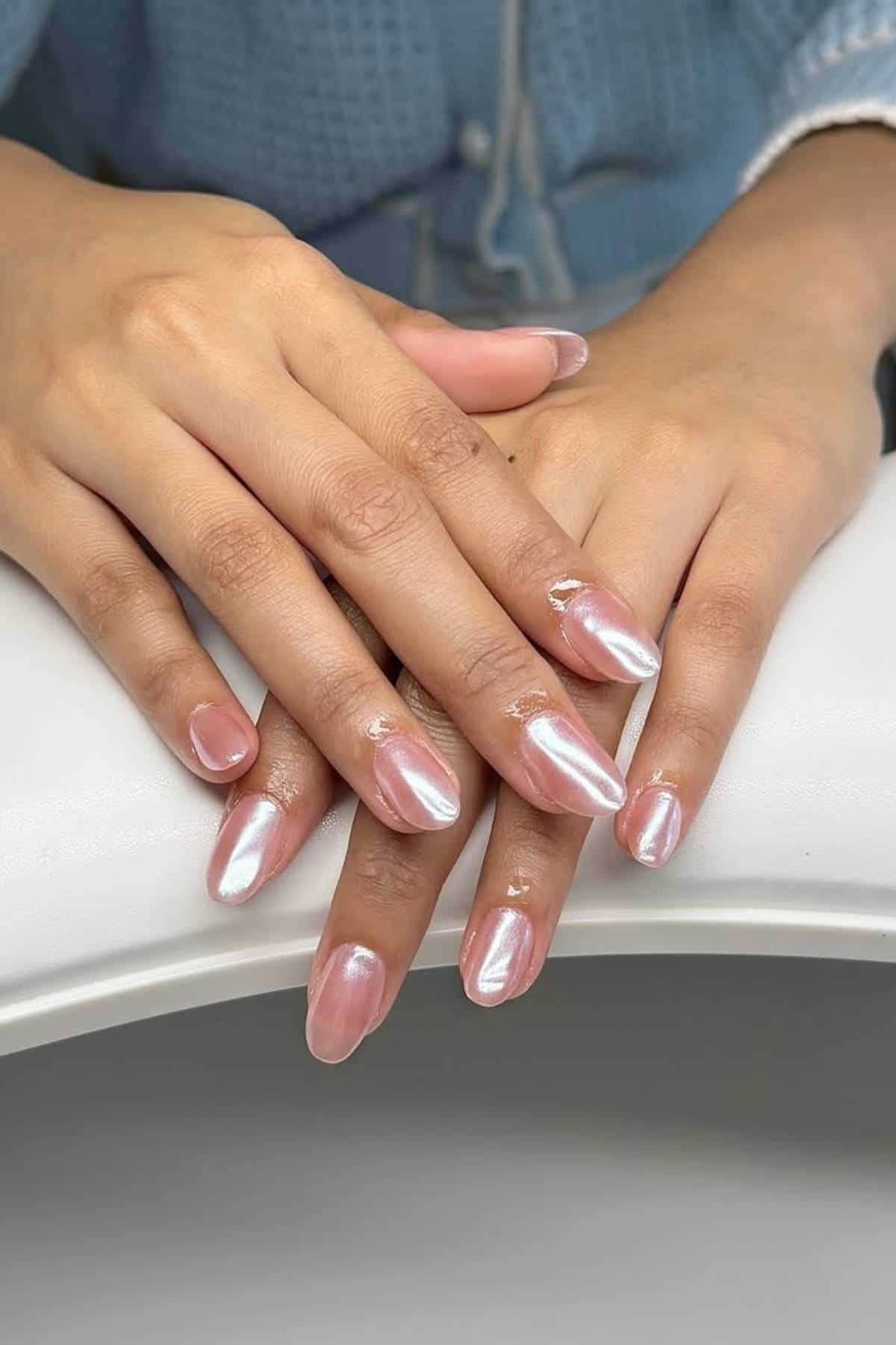 Close-up of two hands with neatly manicured nails painted in a shimmery, light pink polish. The person is wearing a blue textured sweater.