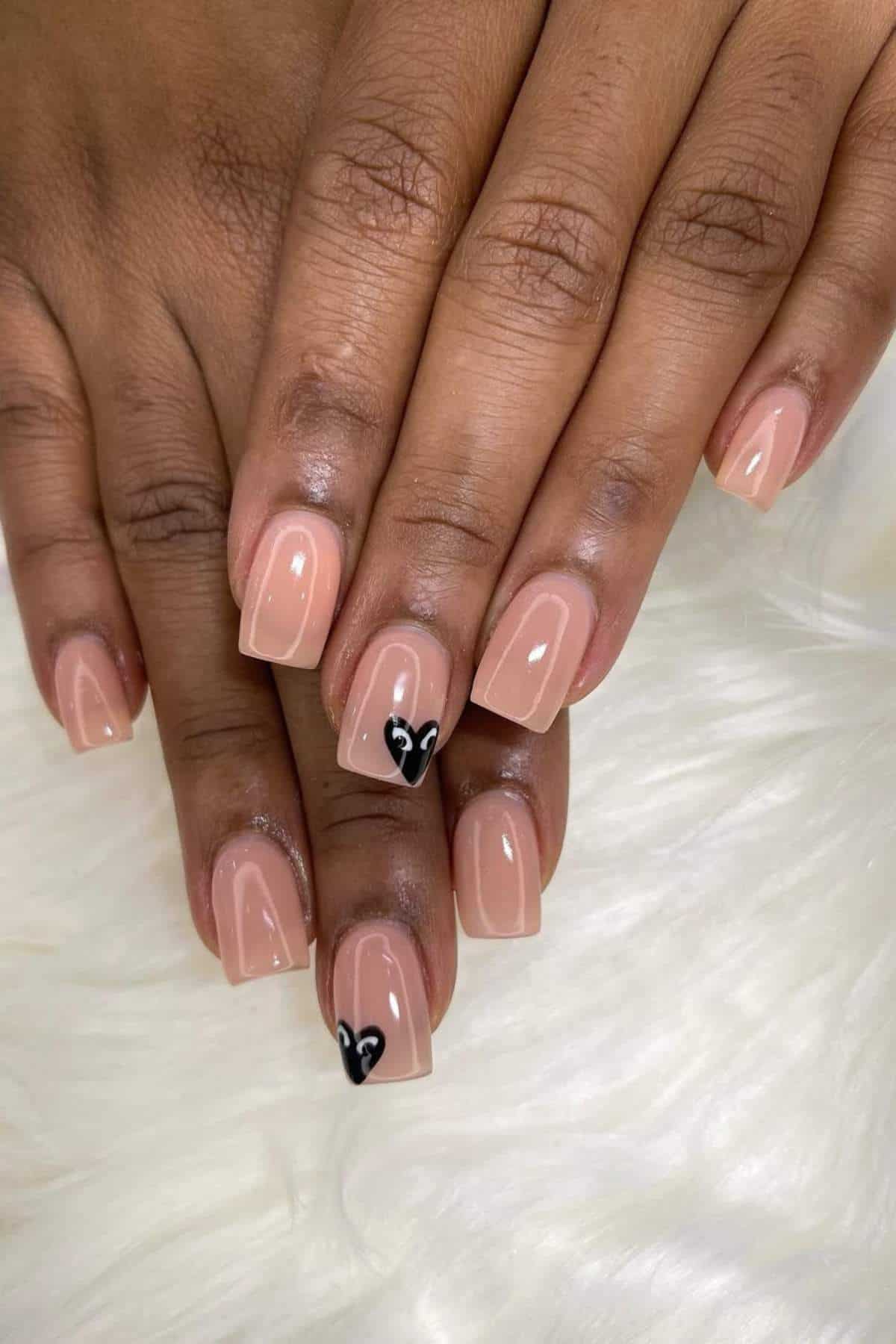 Two hands with short, square-shaped nude pink nails; one nail on each hand has a small black heart design near the tip.