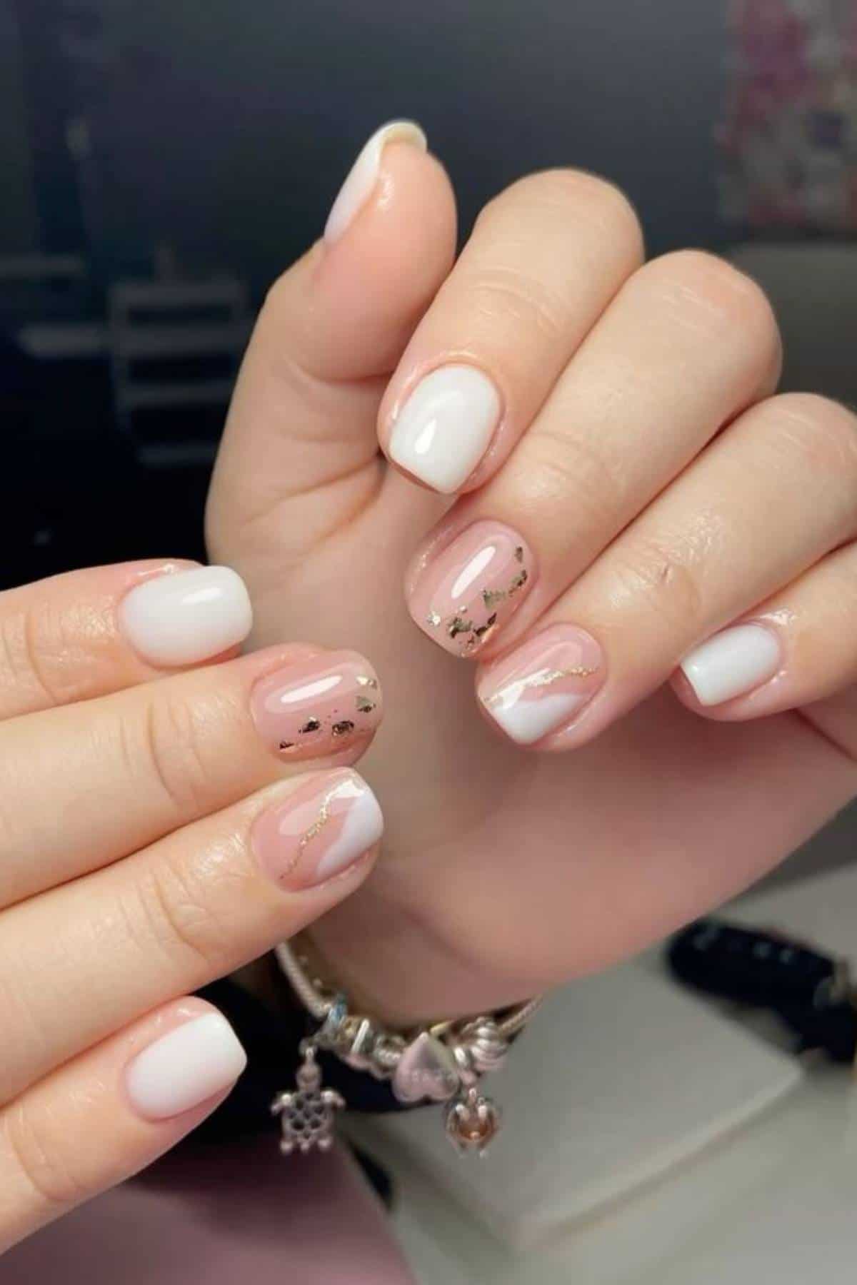 A hand with short, neatly manicured nails featuring a white polish and accent nails with clear polish, metallic flakes, and gold lines.