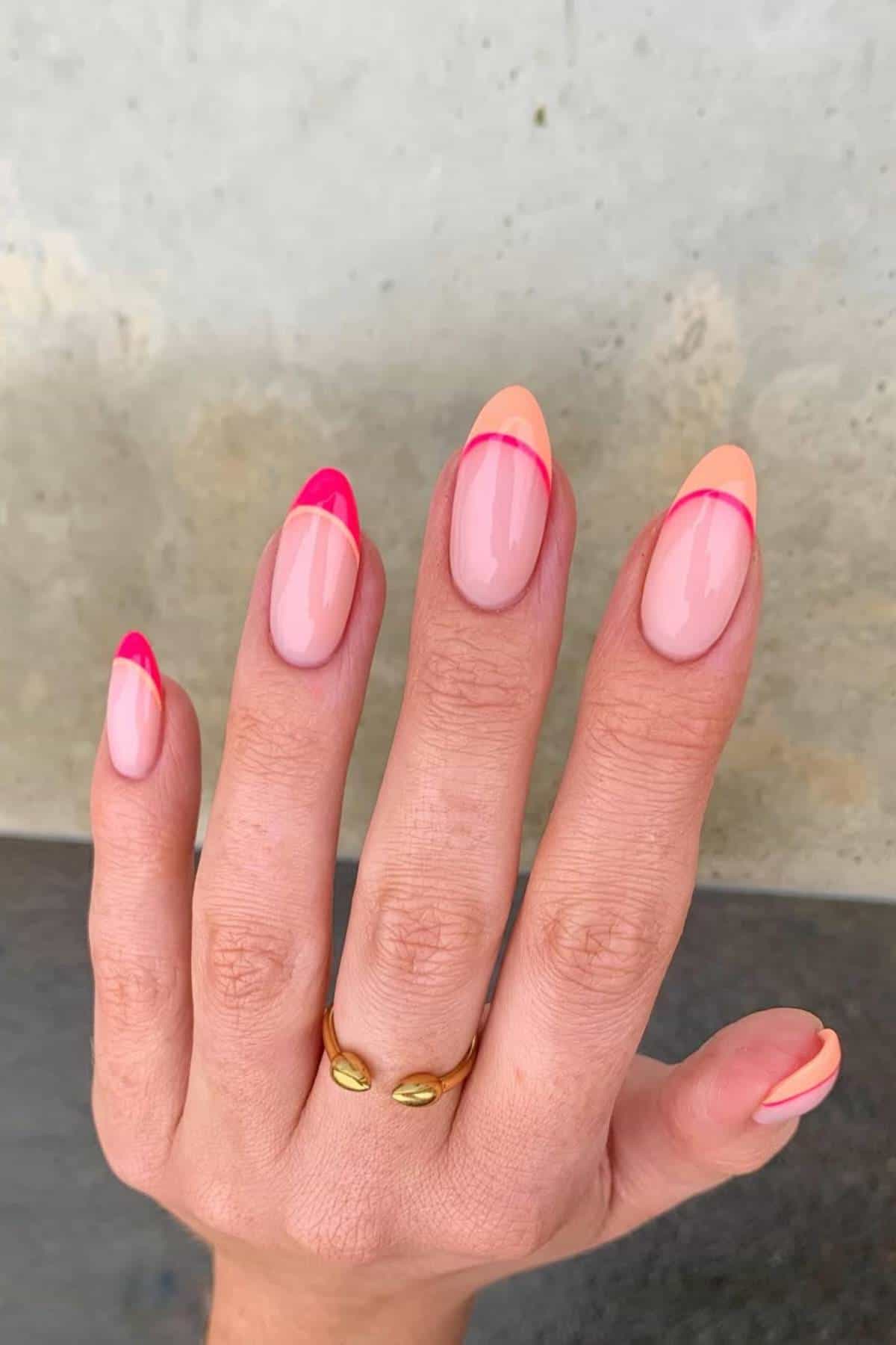 A hand with almond-shaped nails featuring a light pink base and vibrant pink and peach French tips, wearing a gold ring with two oval ends.