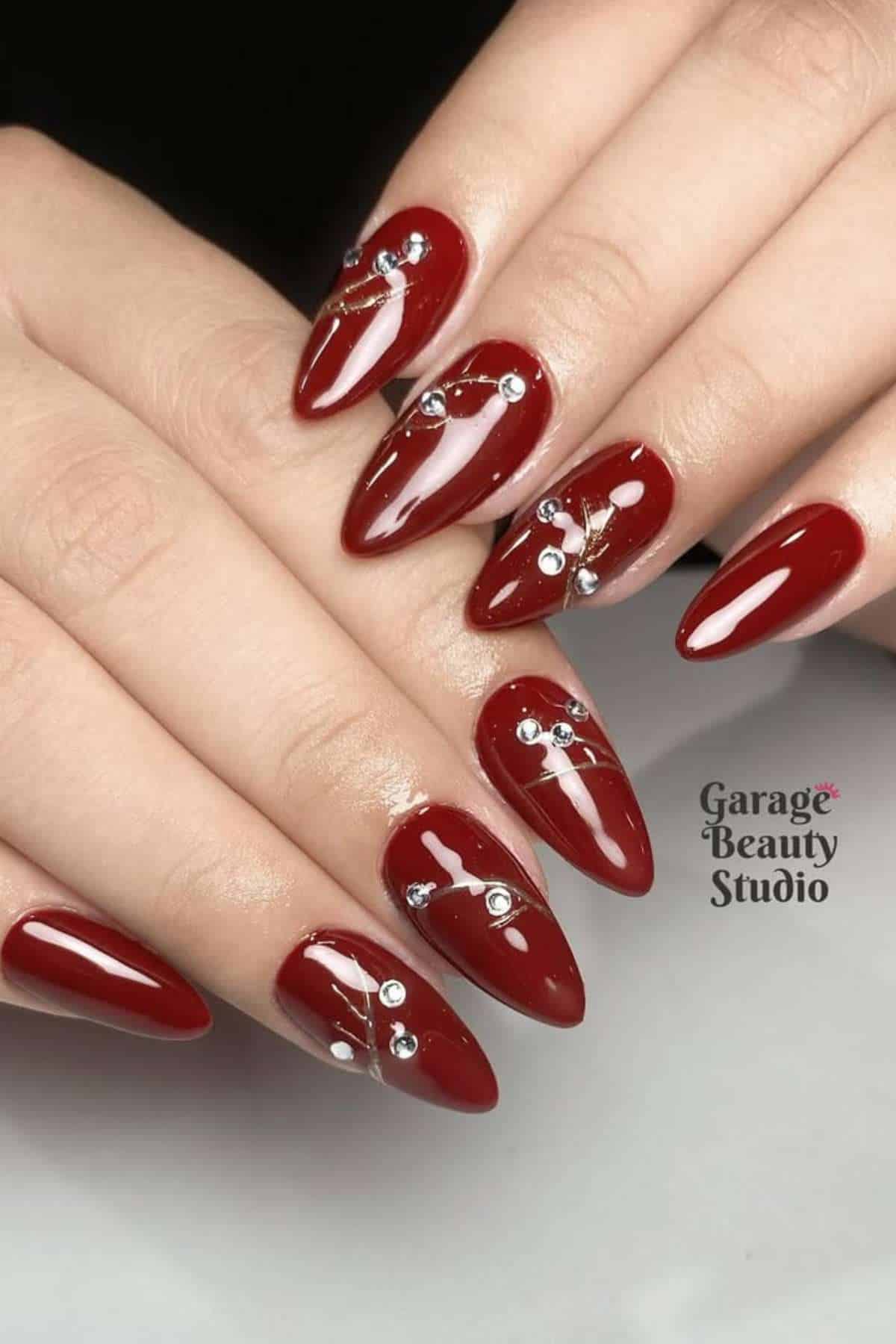 A close-up of hands with long, almond-shaped nails painted dark red and decorated with silver rhinestones and lines.