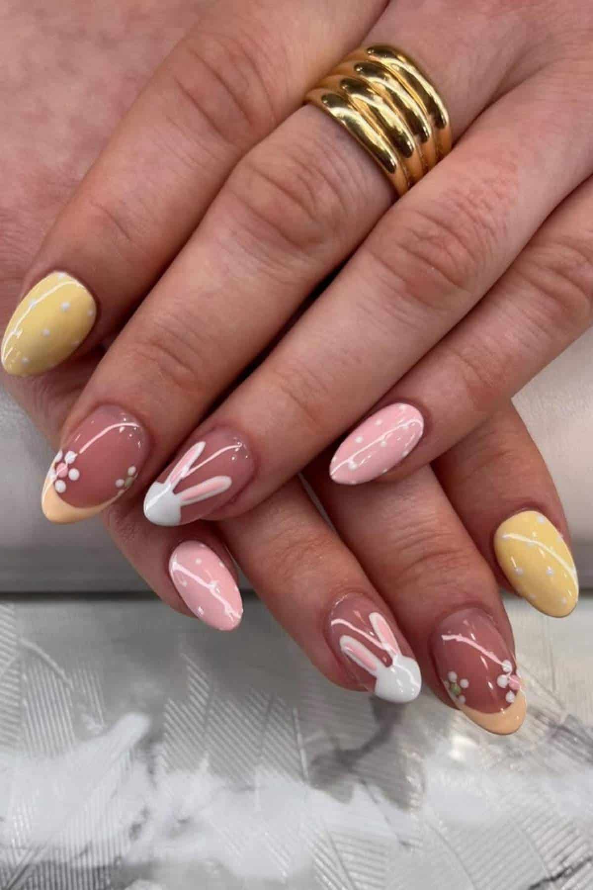 Hands with almond-shaped nails featuring pastel yellow and pink polish, white polka dots, bunny designs, and small white and yellow flower accents. Gold ring on one finger.