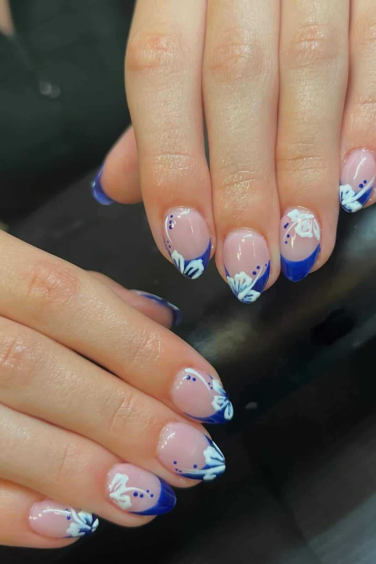 Hands with almond-shaped nails featuring a nude base, blue French tips, and white floral designs with blue accents on each nail.