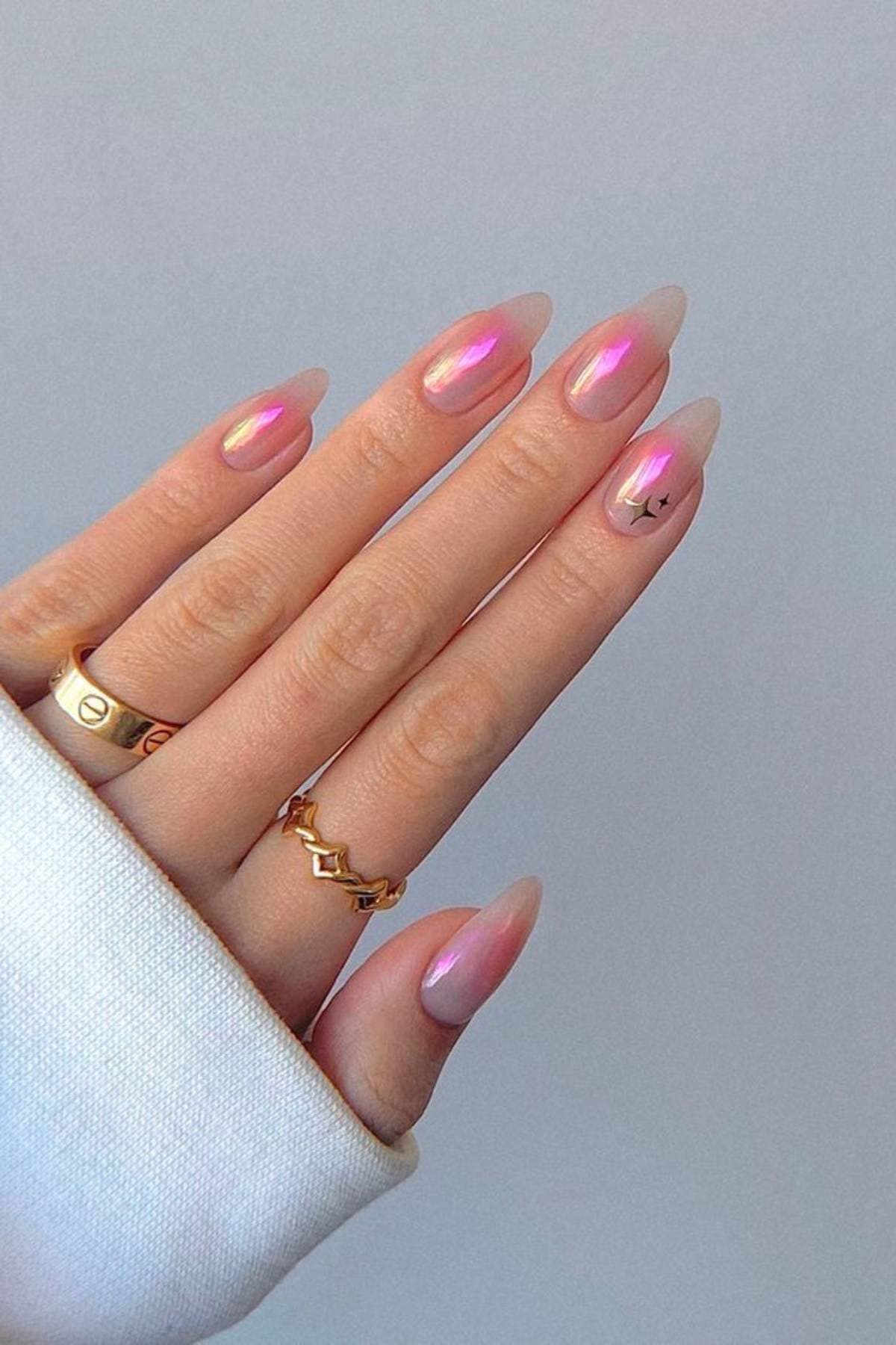 A hand with long, almond-shaped, iridescent pink nails, one with a small black star design, wearing gold rings and a white sleeve.