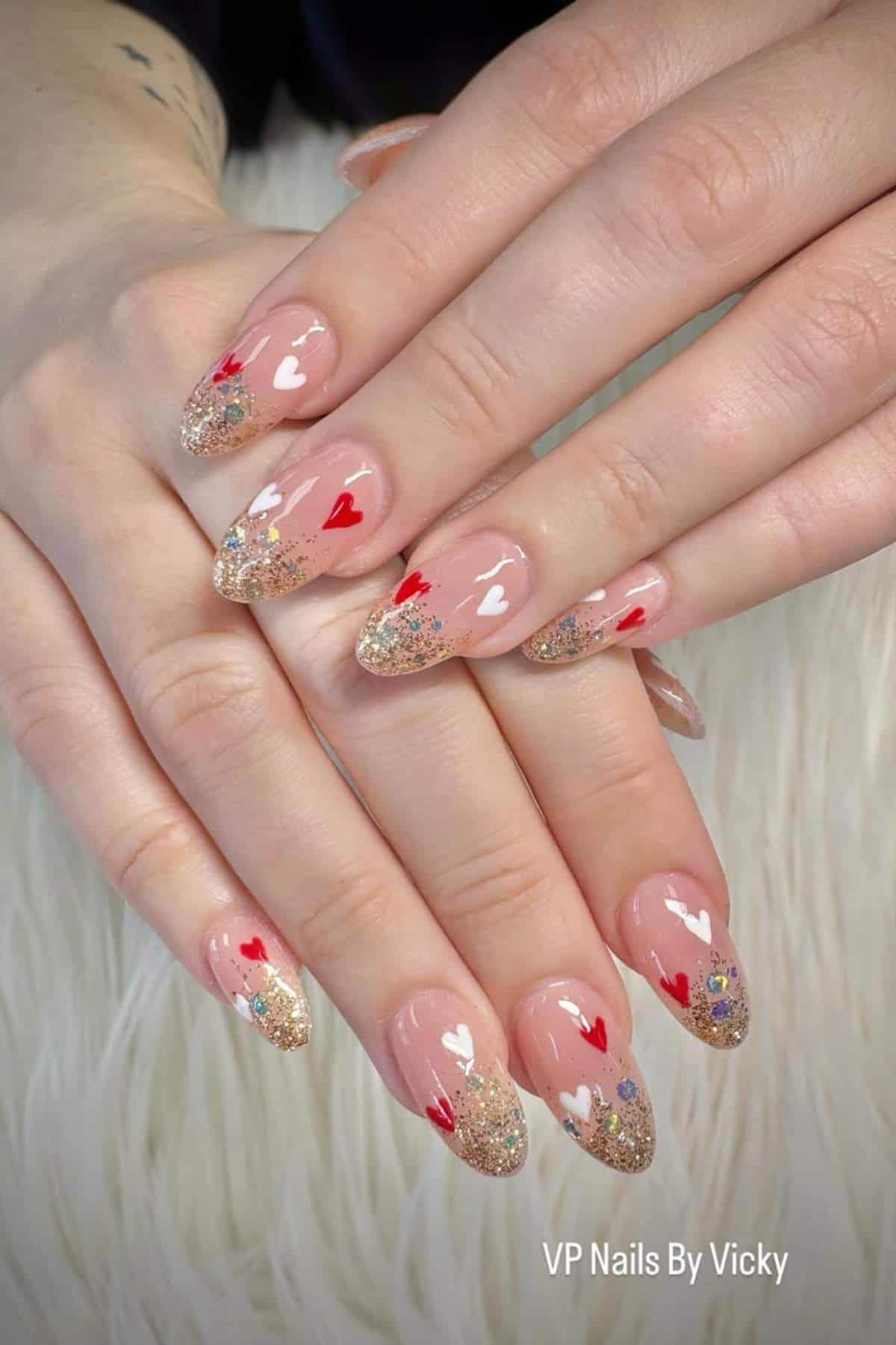 Hands with almond-shaped nails featuring gold glitter tips, red and white heart designs, and small colorful decorations on a soft white surface.