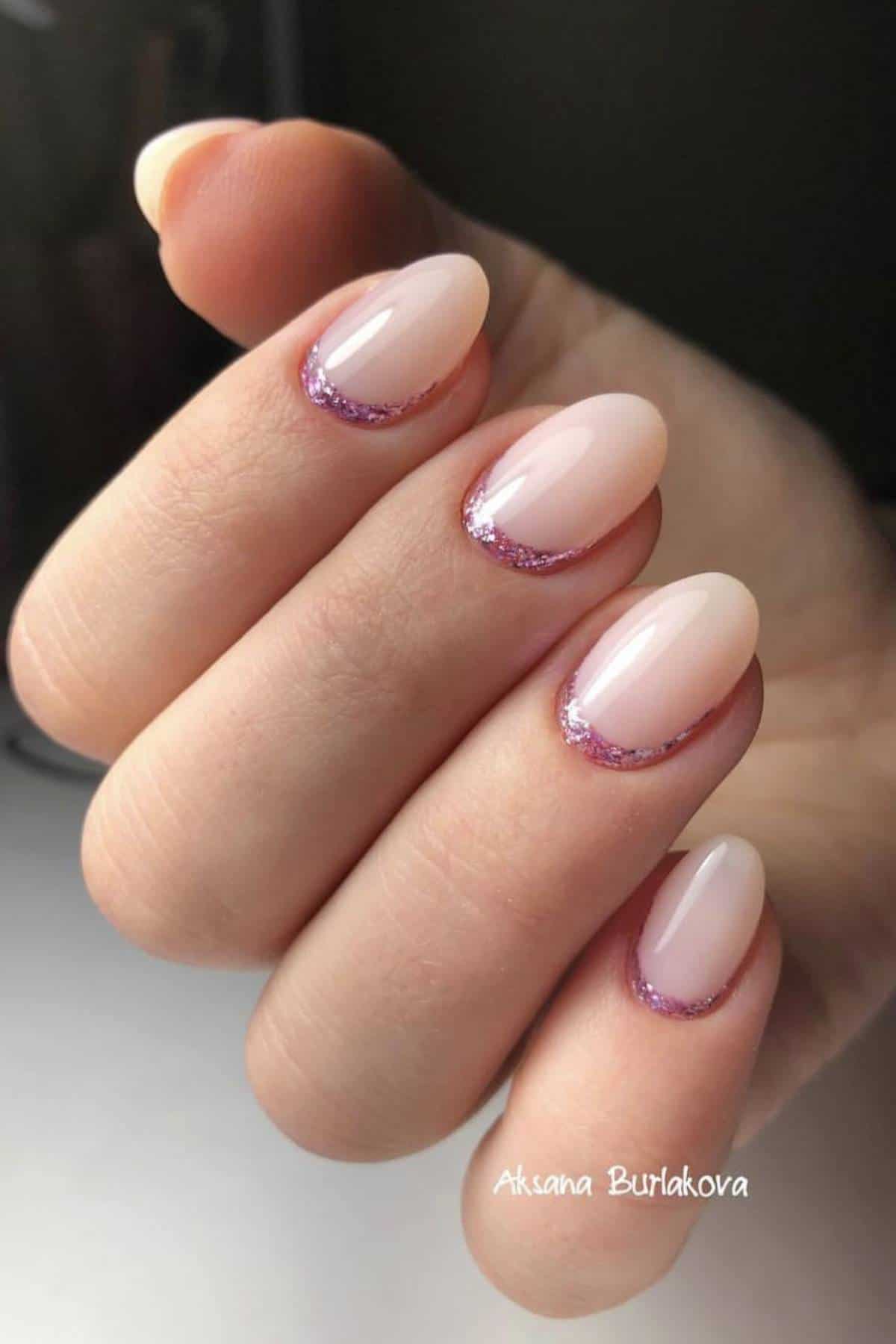 A hand with almond-shaped nails painted in a light nude shade, featuring a subtle pink glitter detail near the cuticles.