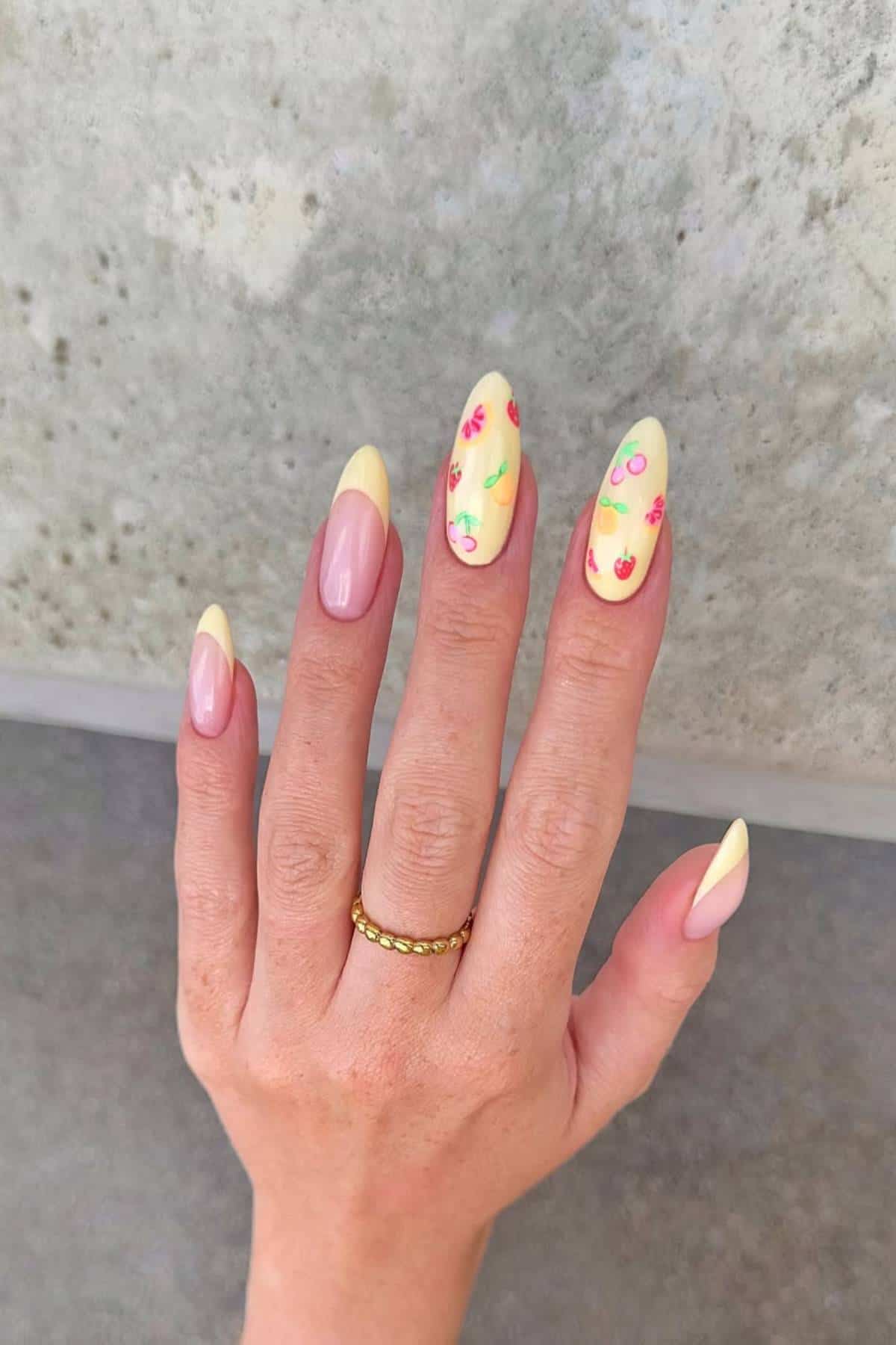 A hand with almond-shaped nails, some painted light pink and others yellow with small floral designs, against a light gray background. The person wears a gold ring.