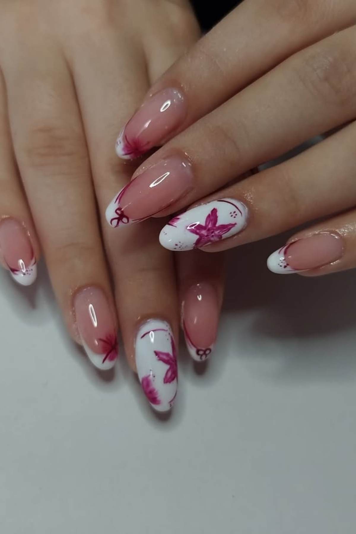 Hands with long, almond-shaped nails featuring a pink and white French tip design with pink floral accents and small decorative elements.