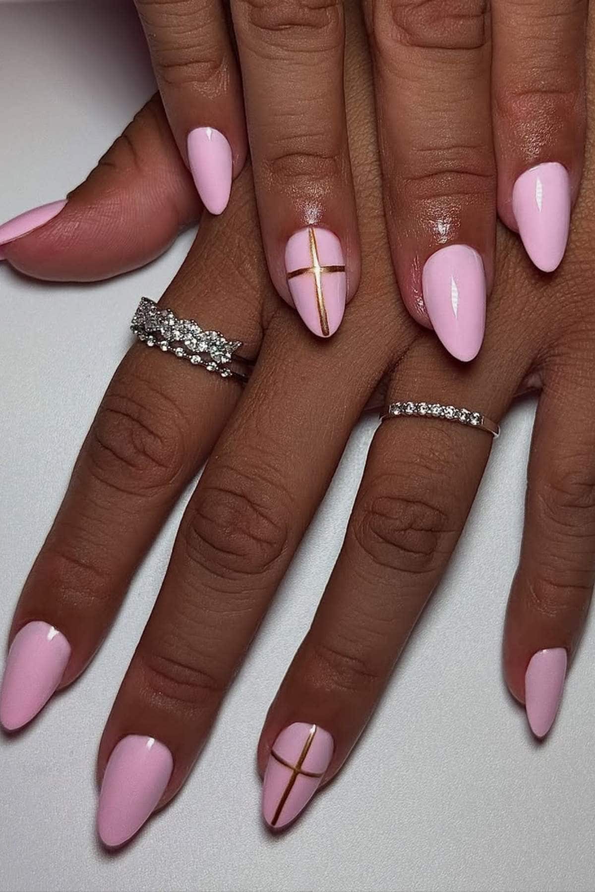 Hands with almond-shaped, light pink polished nails; two nails feature a gold cross design. The person is wearing three silver rings with small stones.
