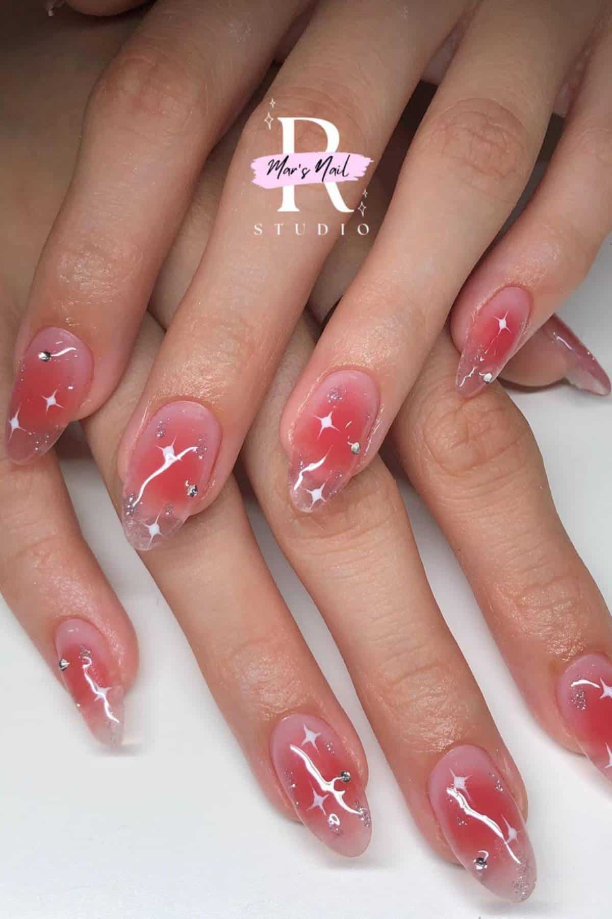 Hands with almond-shaped nails featuring a pink and clear ombre base, decorated with white star designs and small silver gems.