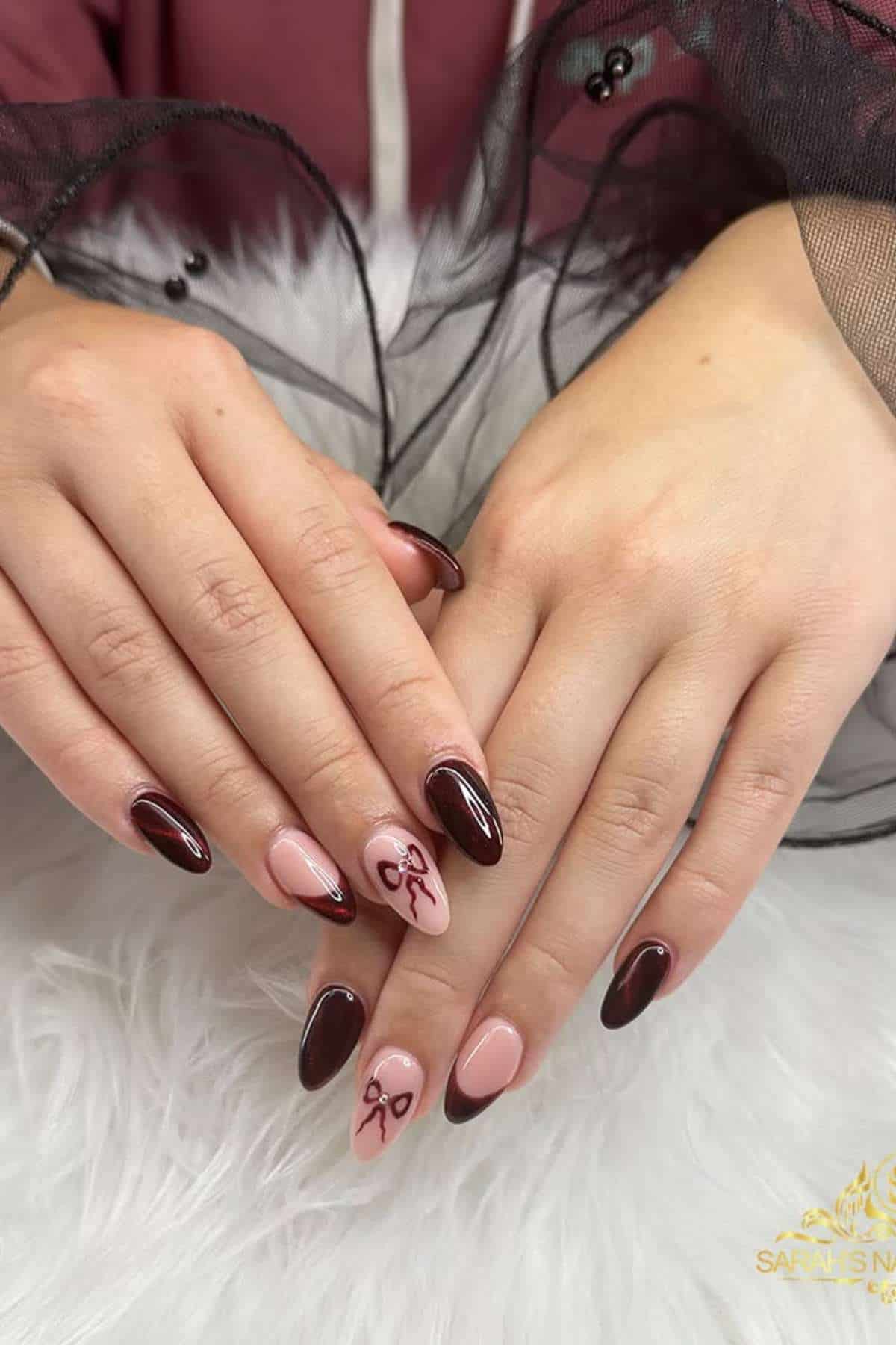 Hands with almond-shaped nails painted in dark burgundy and nude pink polish, featuring small bow designs on the ring fingernails.