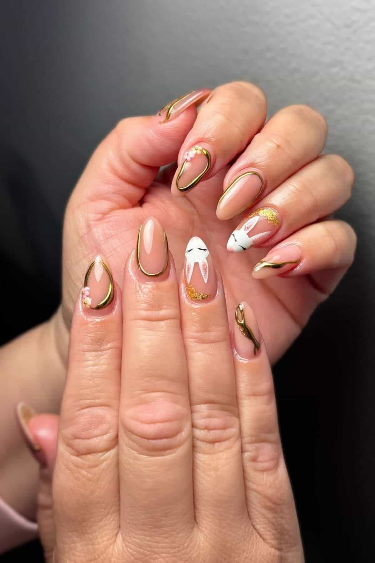 Close-up of hands with almond-shaped nails featuring nude polish, gold accents, and an Easter bunny design on the middle finger.