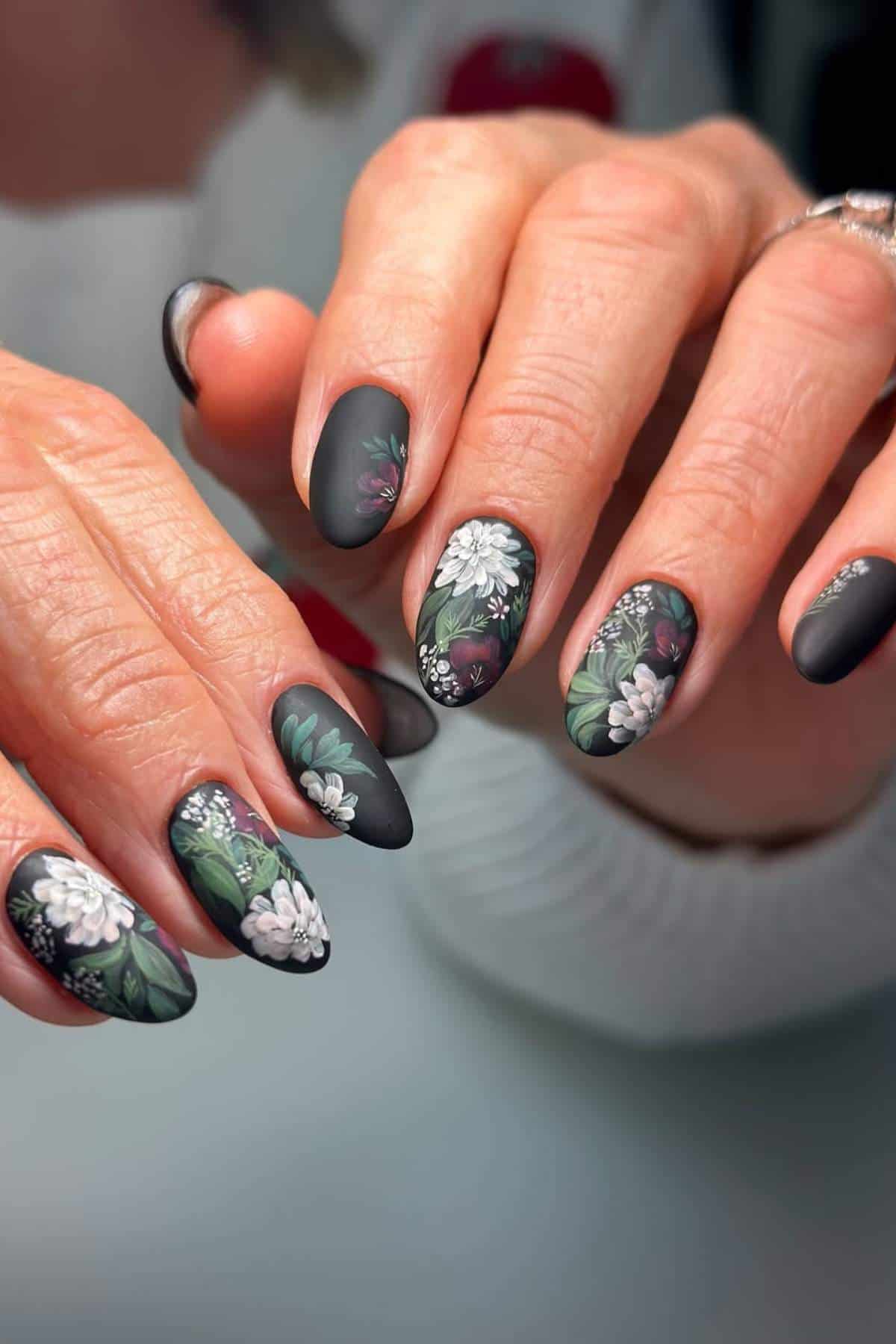 Close-up of hands with long, almond-shaped nails painted matte black and decorated with detailed floral designs in white, green, and purple.