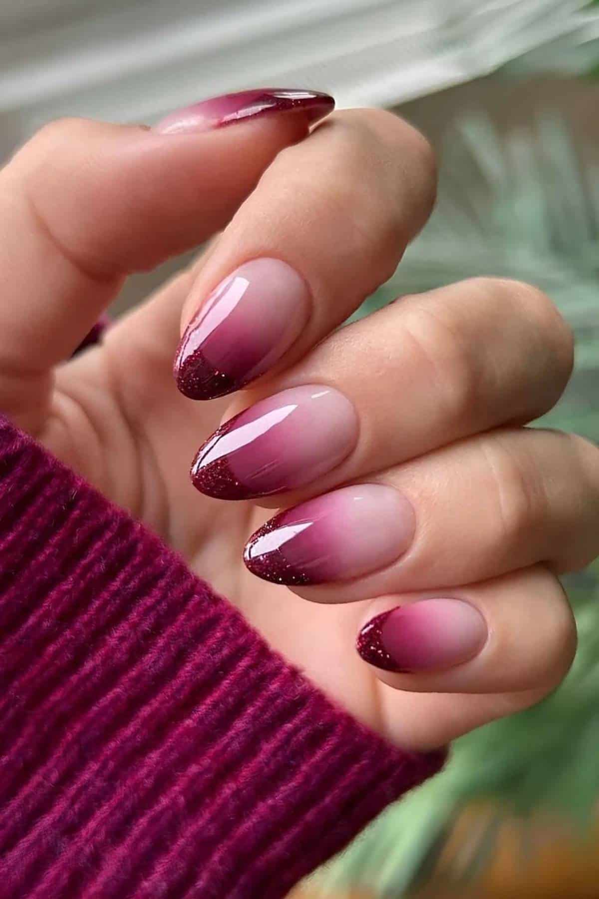 A hand with almond-shaped nails featuring a glossy burgundy and nude ombre design with glittery burgundy tips, shown against a blurred background.