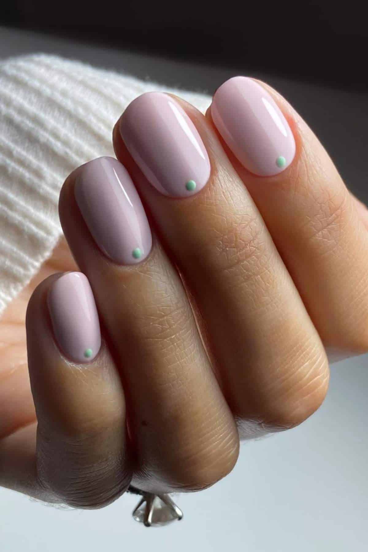 A hand with short, rounded nails painted light pink, each featuring a small green dot near the cuticle.