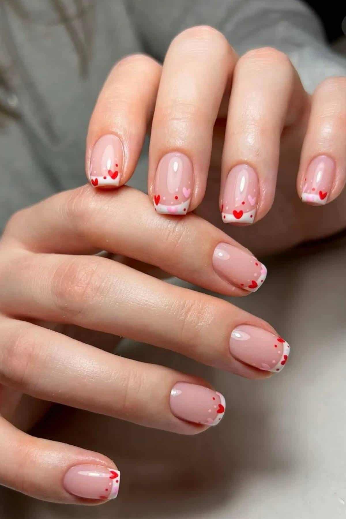 Close-up of hands with manicured nails featuring a French tip design accented with small red hearts on a nude base.