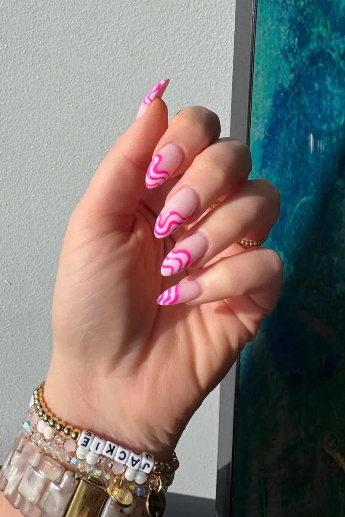 A hand with long, almond-shaped nails featuring pink and white swirl nail art; the wrist is adorned with multiple beaded and gold bracelets, one spelling "JACKIE.