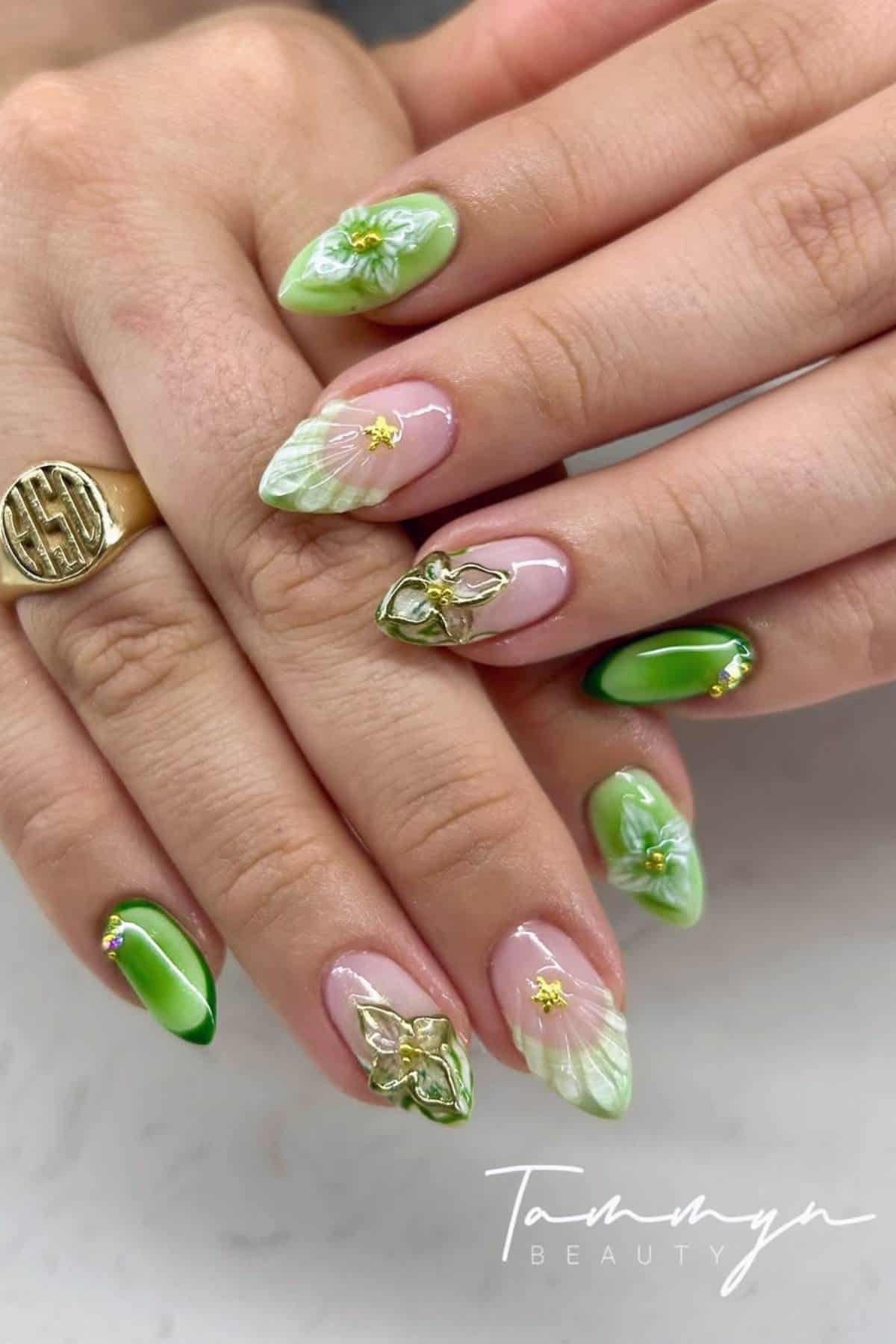 A hand with long almond-shaped nails, decorated with green and white floral designs, gold accents, and 3D embellishments, resting on another hand.