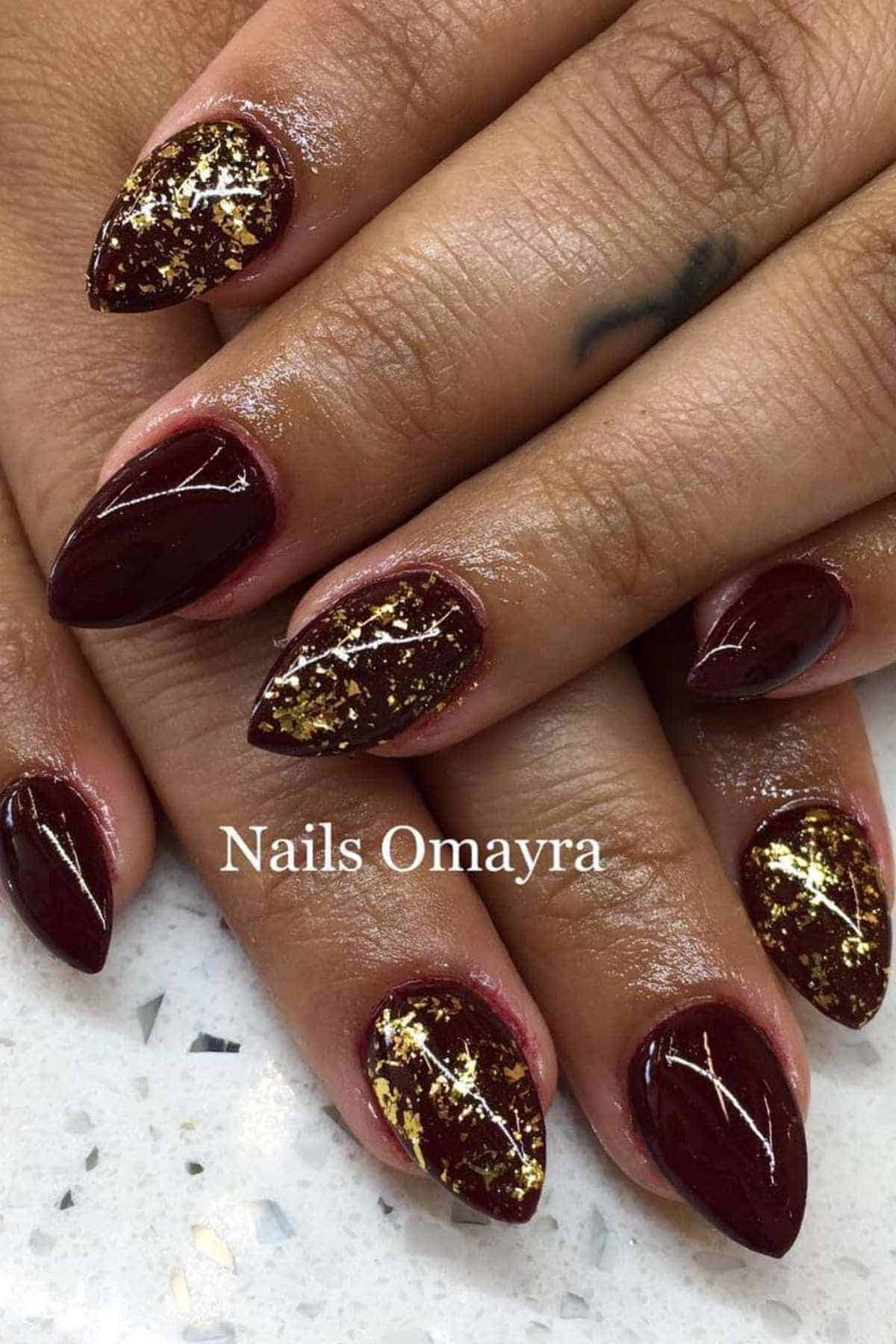 Close-up of hands with almond-shaped burgundy nails; accent nails feature gold foil details. The text "Nails Omayra" appears on the image.