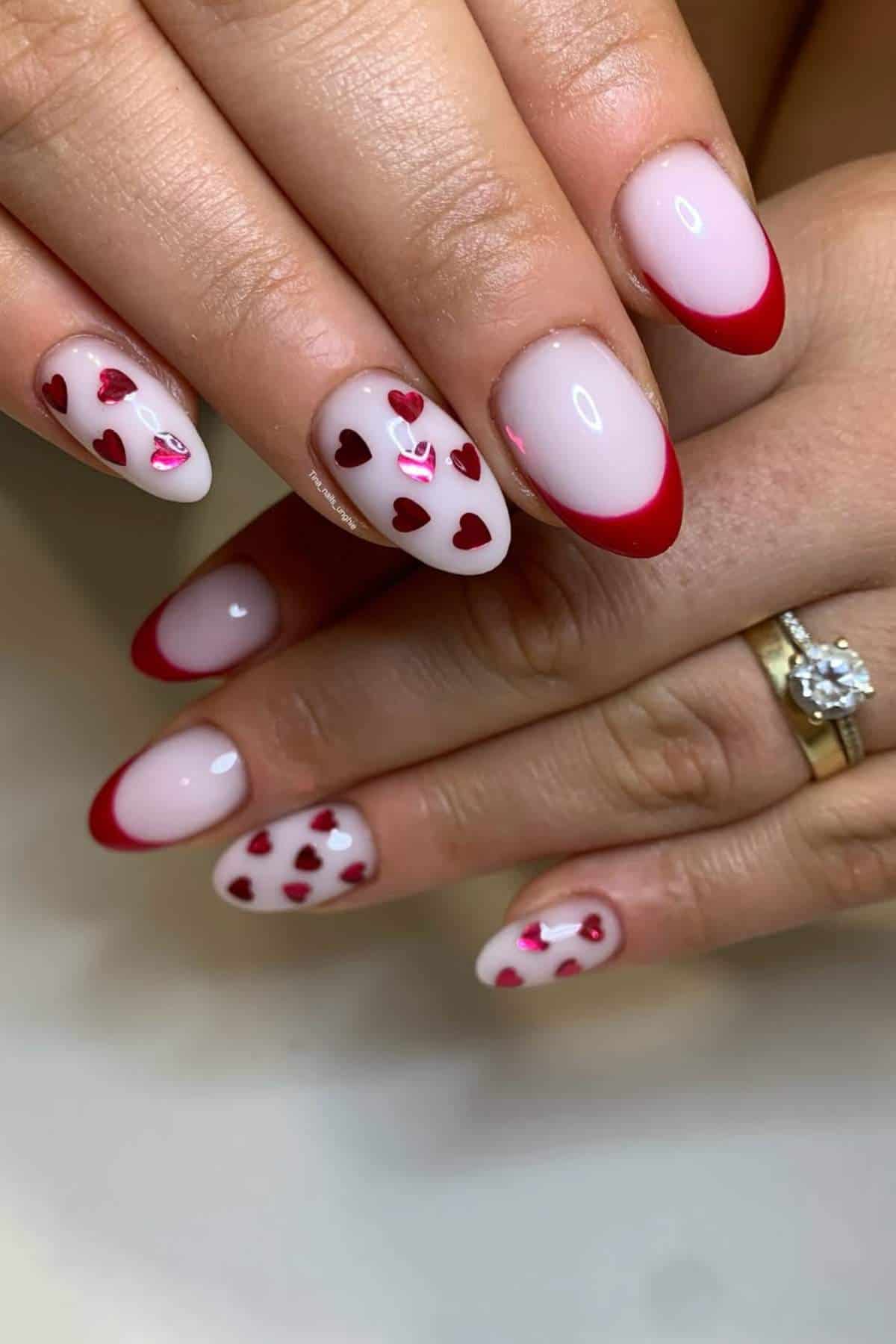 Hands with almond-shaped nails featuring a pink base, red tips, and red heart designs; one hand has a gold ring with a diamond.