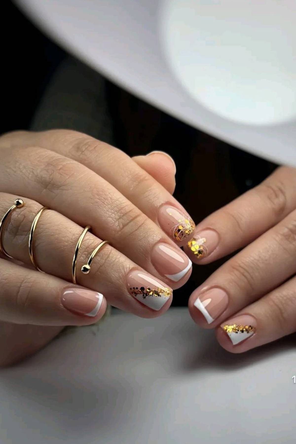 Hands with a French manicure featuring white tips, gold glitter accents, and geometric designs on some nails. The person is wearing several gold rings.
