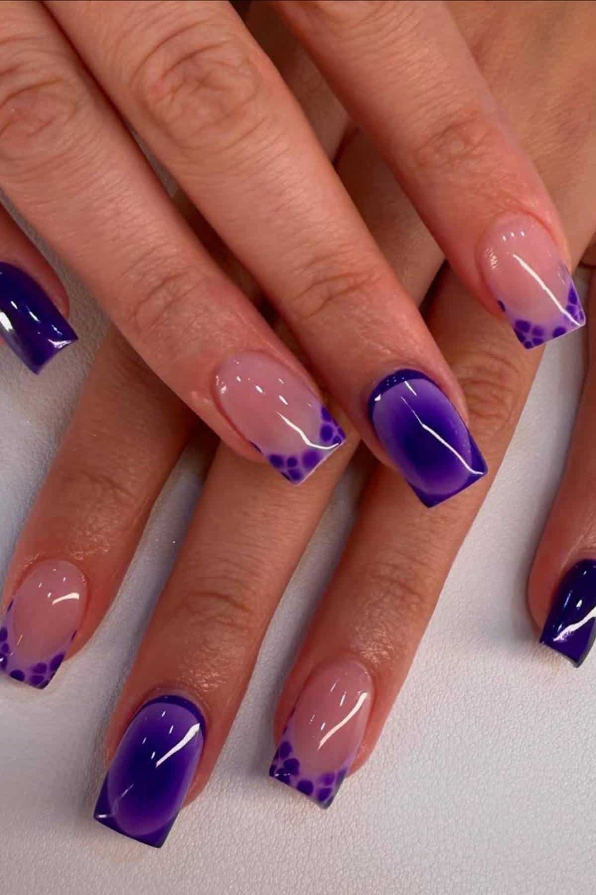 Hands with square-shaped nails featuring a purple gradient French tip design and small purple dots at the corners on a light background.