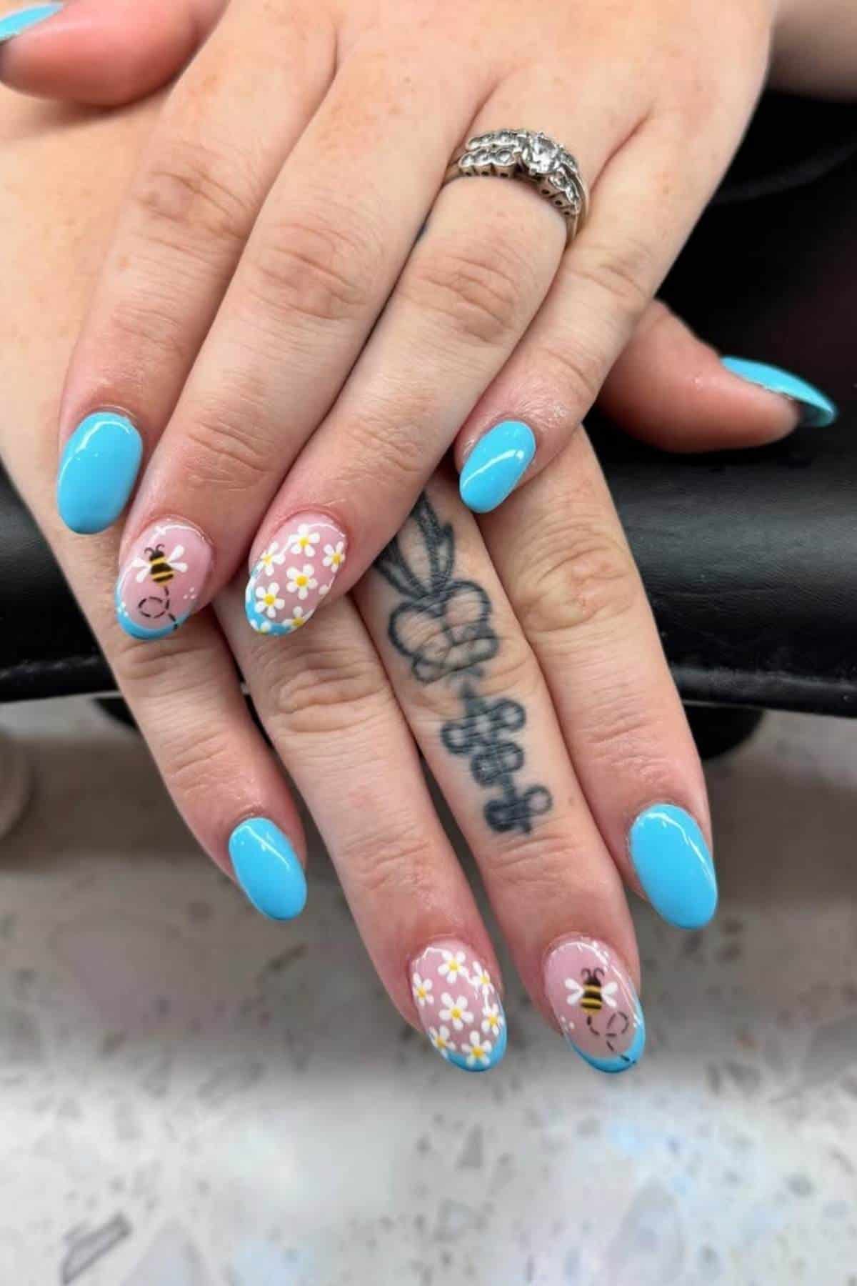 Hands with blue and floral nail art; ring and visible tattoo on fingers. The floral designs feature white and yellow flowers on a nude base with blue tips.