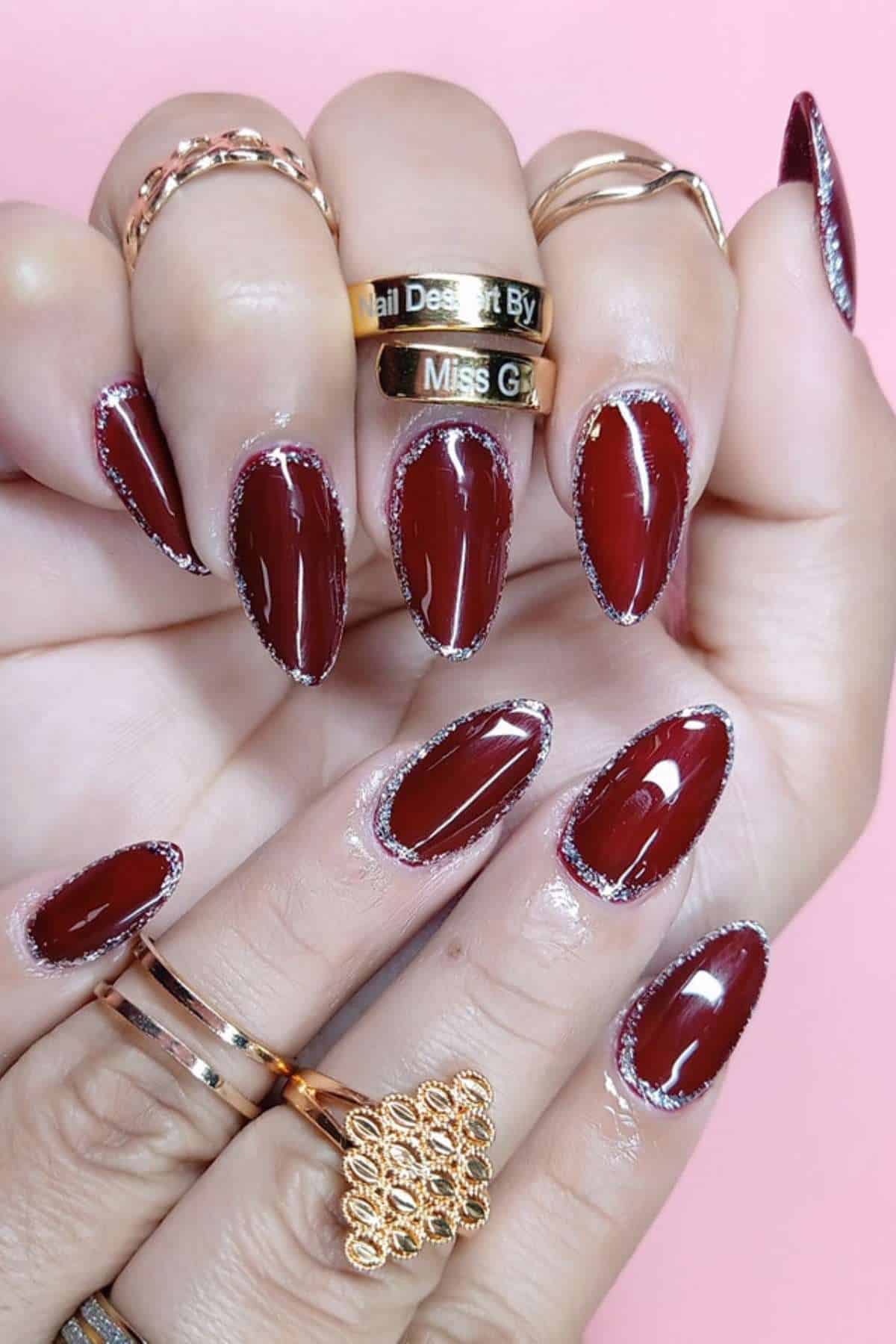 Hands with dark red almond-shaped nails outlined with silver glitter, wearing multiple gold rings, shown against a pink background.