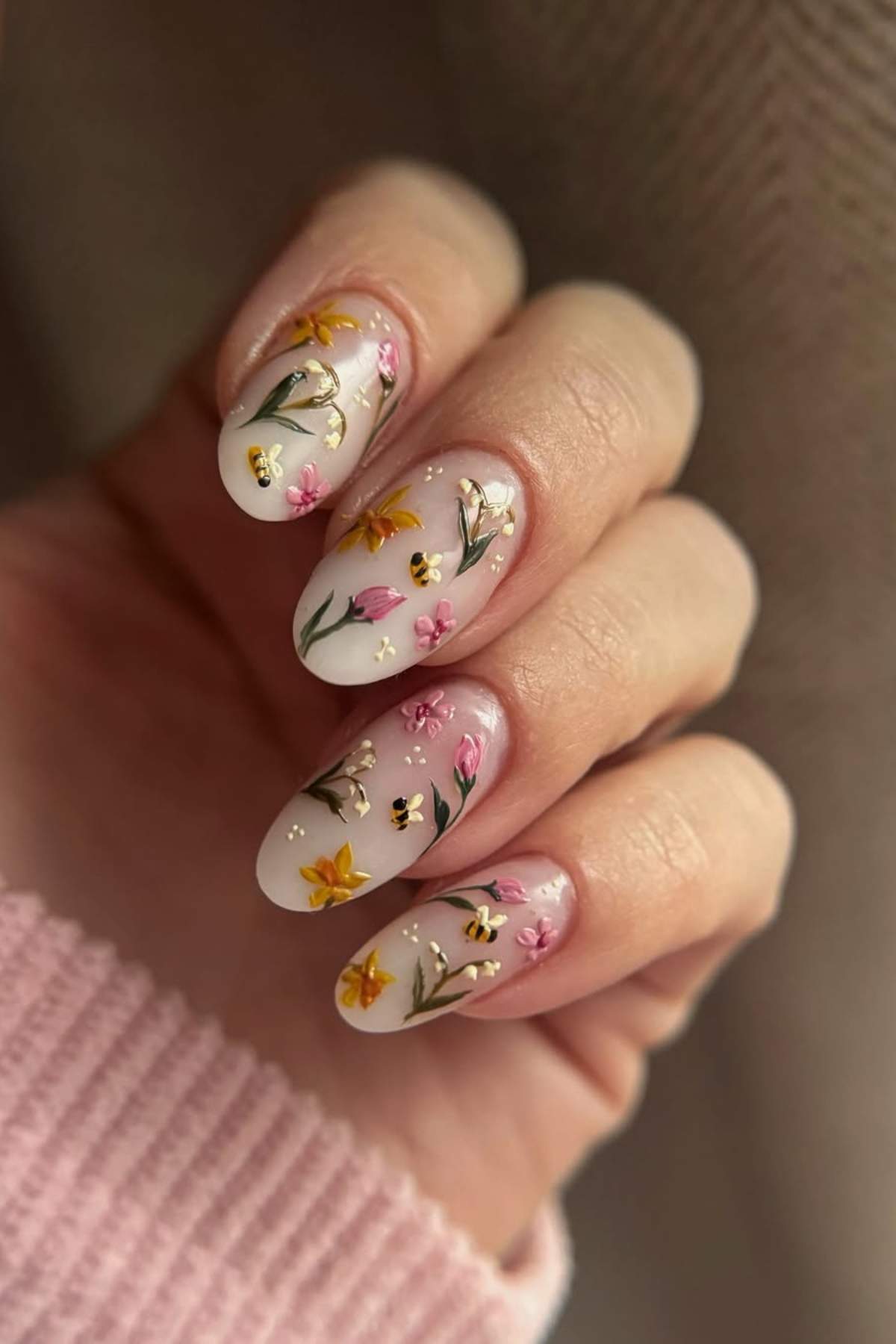 A hand with long, oval-shaped nails painted with a pale base and decorated with detailed yellow, pink, and purple flowers and small bee designs.