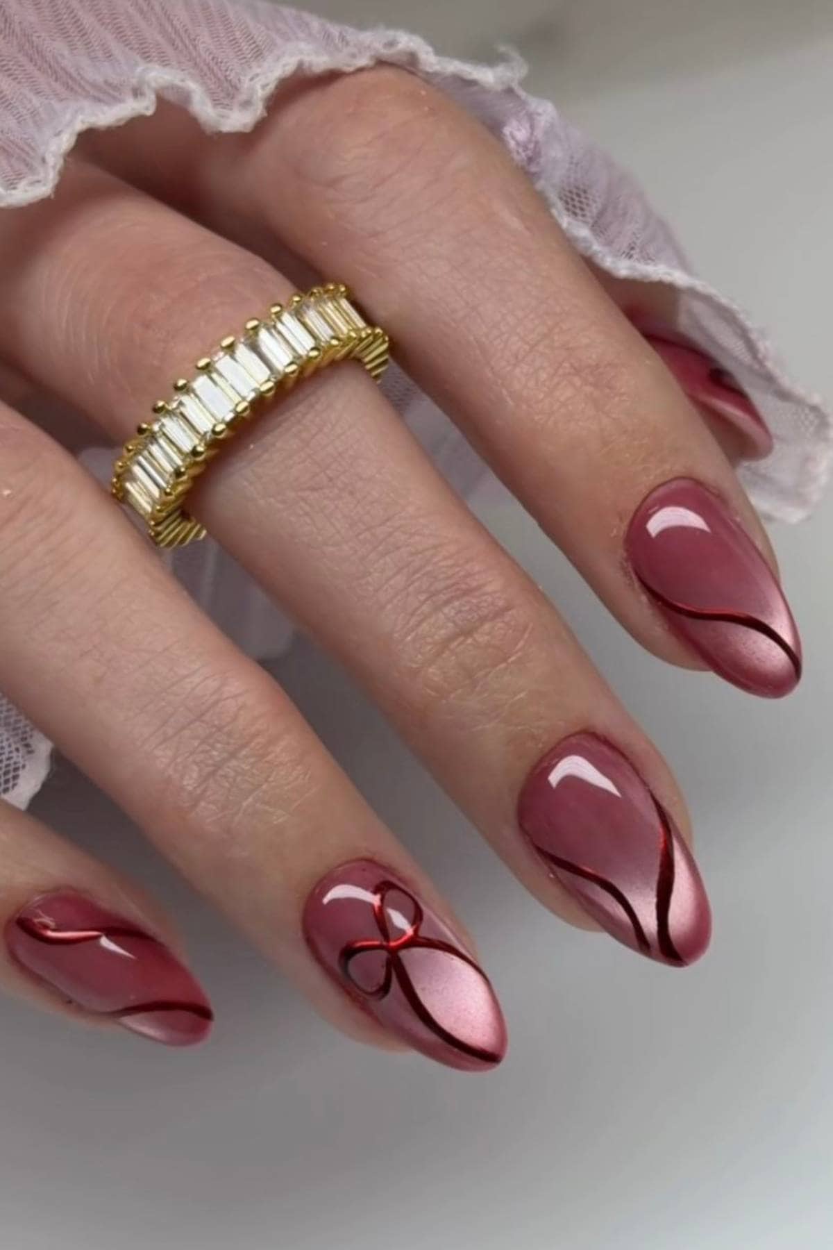 A hand with almond-shaped nails painted in shiny mauve with black and red abstract lines, one nail features a small red ribbon design. The hand wears a gold ring with rectangular stones.