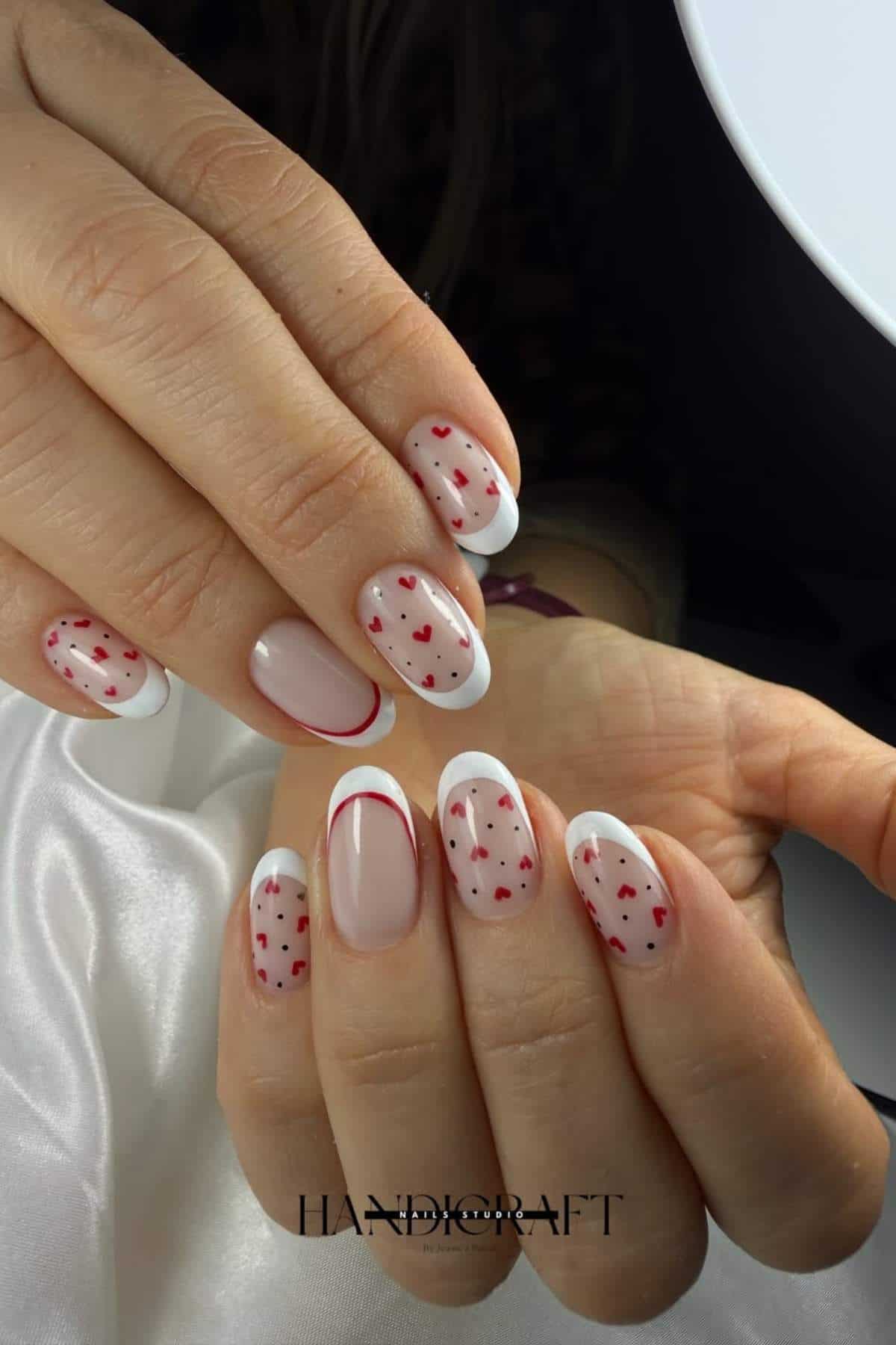 Hands with oval-shaped nails featuring a French manicure and small red heart designs on a nude base.