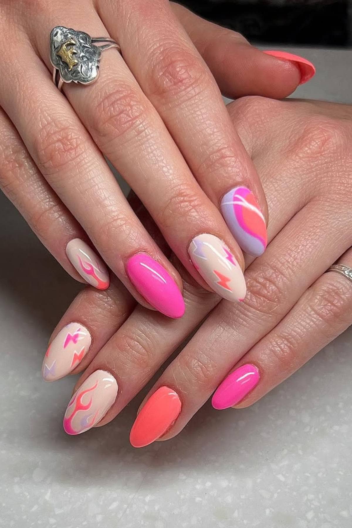 Hands with long almond-shaped nails feature a mix of pink, neon orange, and cream polish with abstract and wavy line designs. The person wears two silver rings.