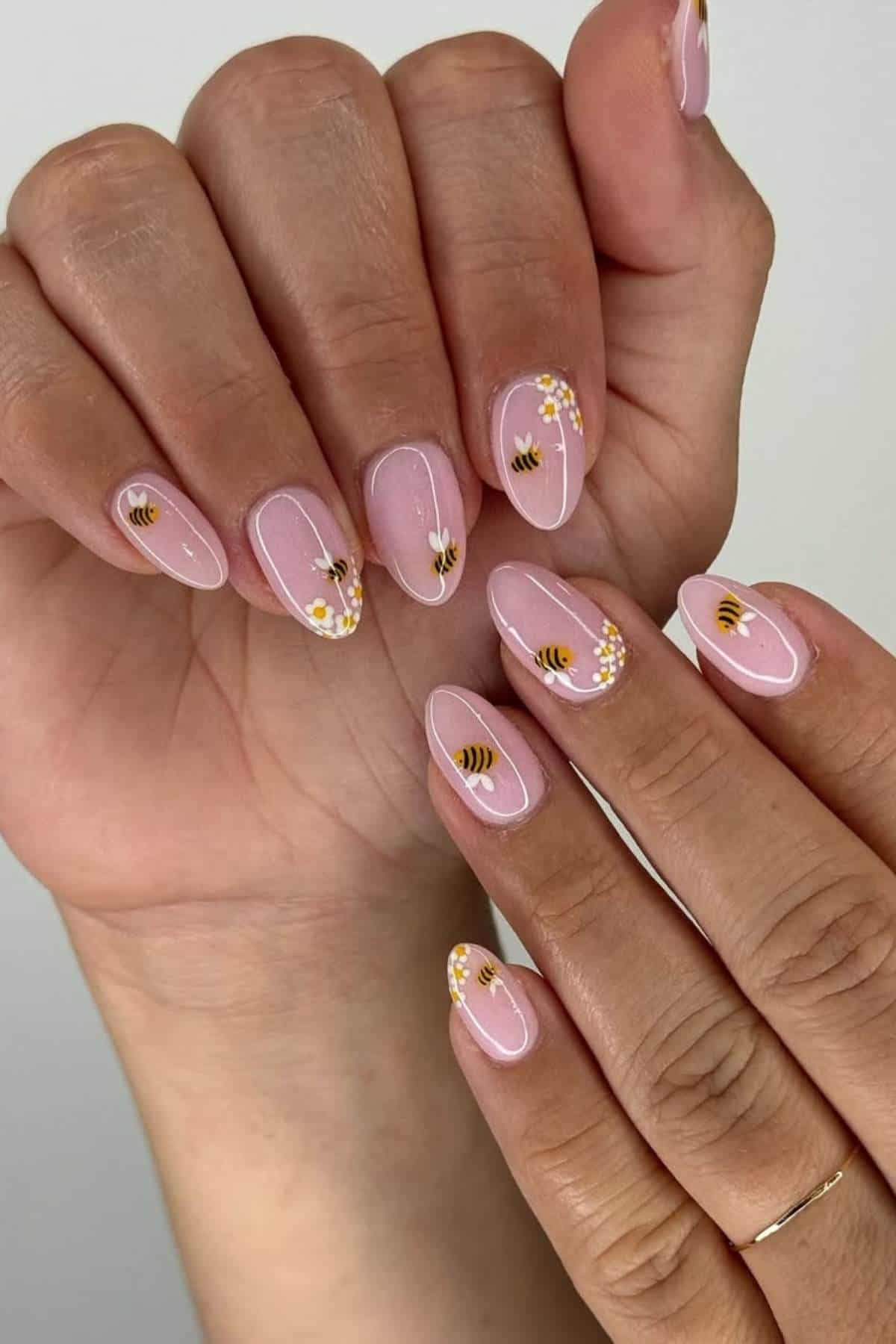 Hands with almond-shaped nails painted pale pink, decorated with small bee and daisy flower designs on each nail.
