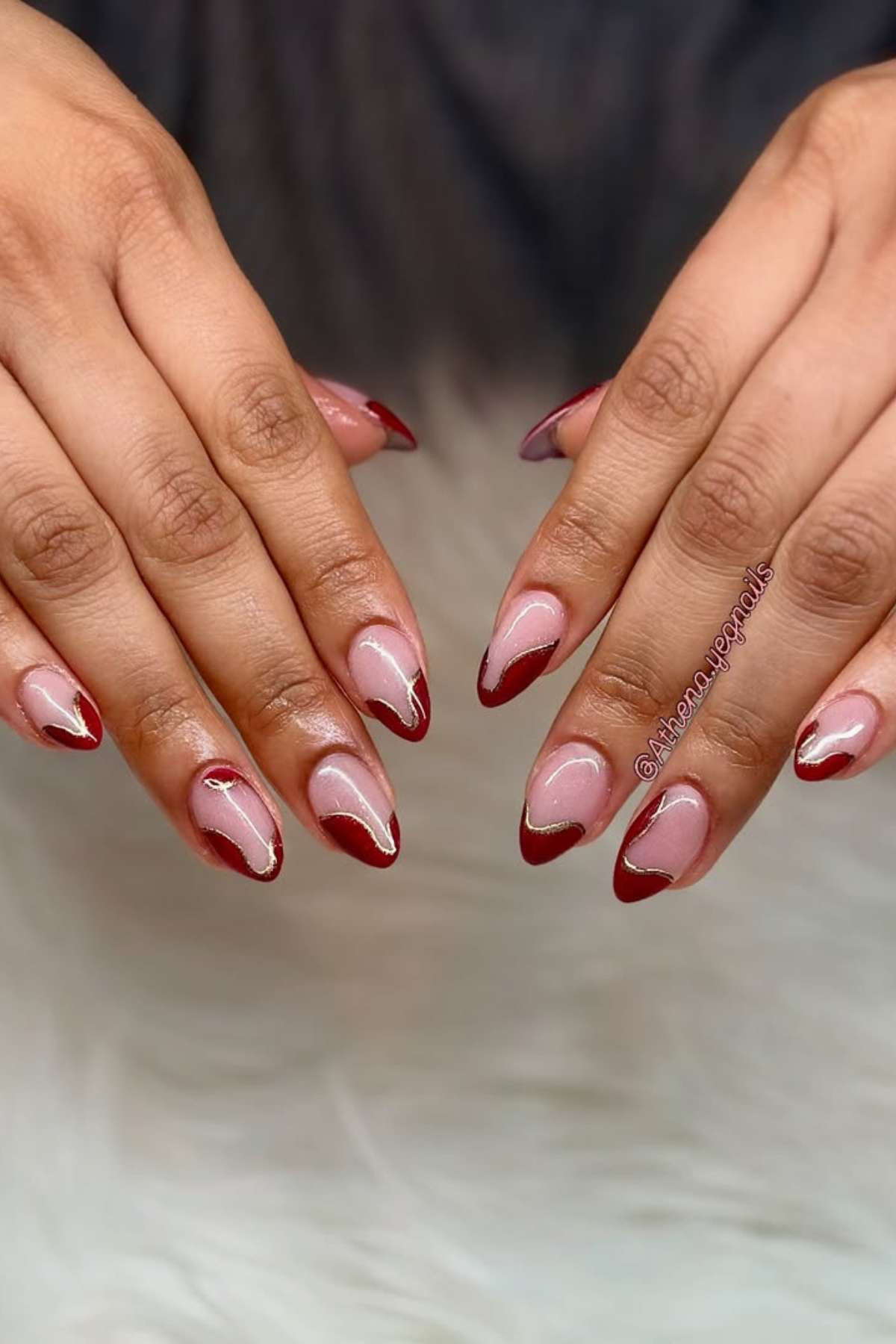 Two hands with almond-shaped nails painted in a nude base color and deep red tips, featuring a thin gold accent line along the tip edges.