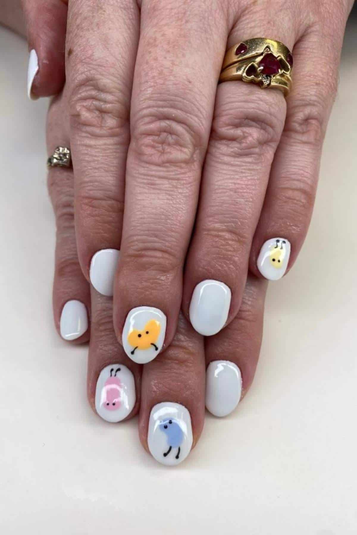 Hands with white painted nails featuring small, colorful designs of animal faces; person is wearing two gold rings, one with a red stone.