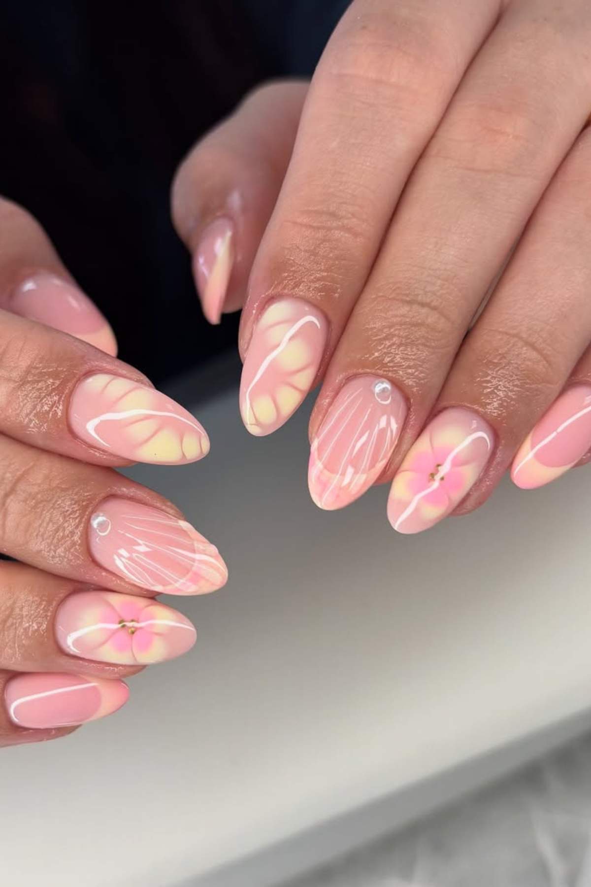 Hands with almond-shaped nails featuring a pink and yellow abstract floral design, white accents, and small rhinestone embellishments.