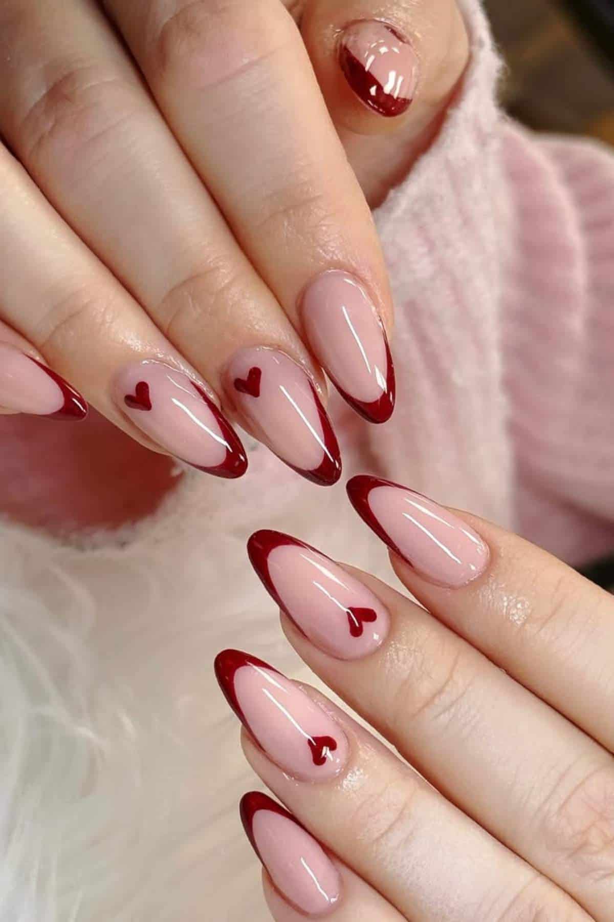 Hands with almond-shaped nails featuring a nude base, deep red French tips, and small red heart designs on some nails. The person is wearing a pale pink sweater.