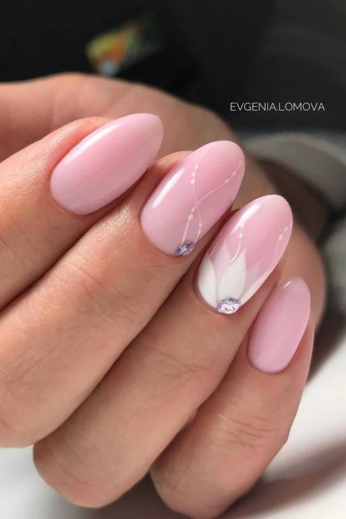 A hand with oval-shaped, light pink gel nails; two nails feature white floral designs and small rhinestone accents.