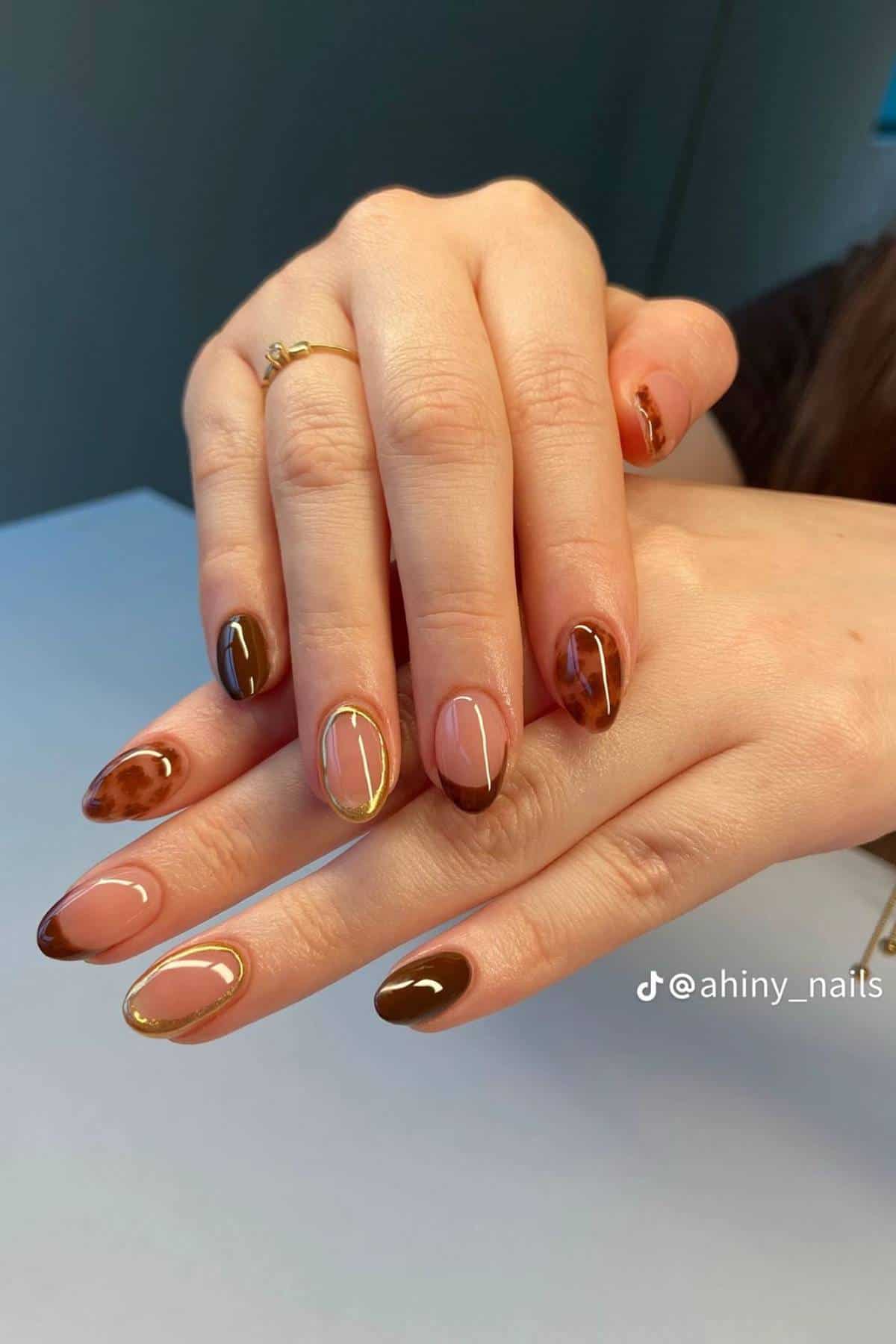 A hand with almond-shaped nails features a brown and gold abstract nail art design on a light background.